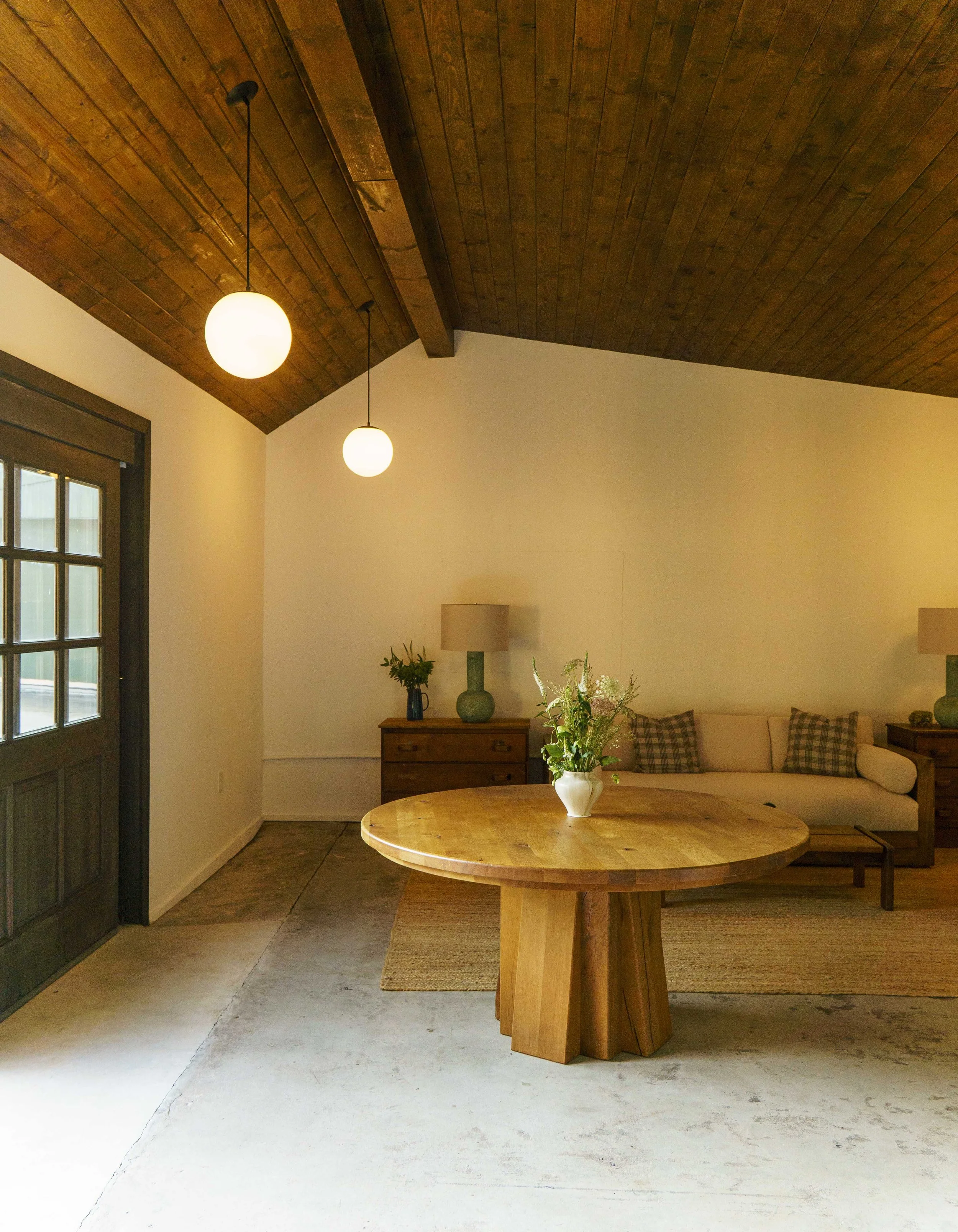 Living room with a wooden ceiling, hanging globe pendant lights, a window with wooden trim, a sofa with plaid pillows, a wooden sideboard with a green vase and plants, a round wooden table with a white vase and flowers, beige carpet, and a floor lamp.