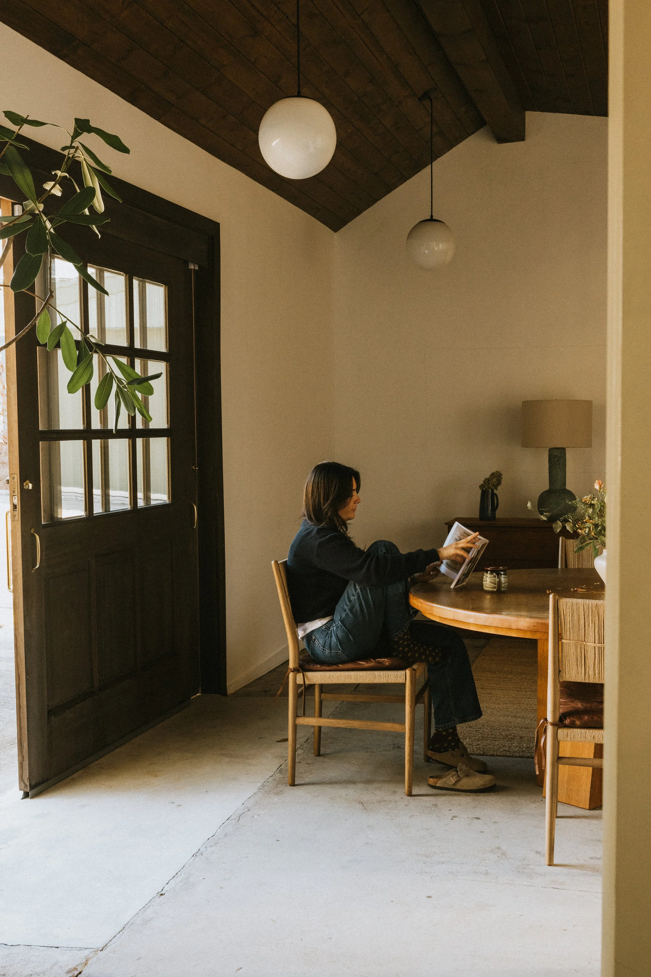 A woman sitting at a wooden dining table reading a book or magazine in a cozy, well-lit room with a dark wood ceiling, wall-mounted lamp, and decorative plants.