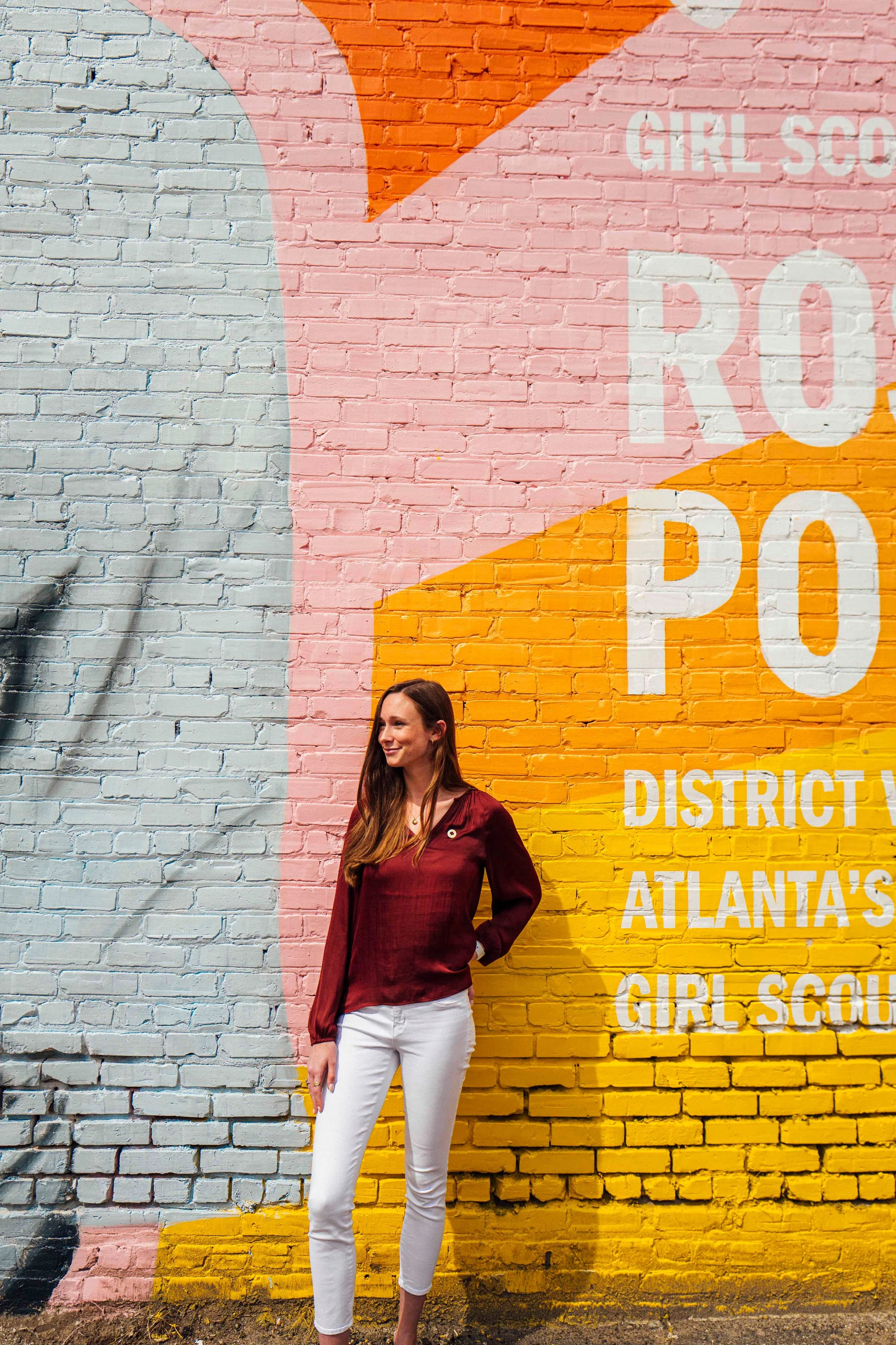 Woman with long brown hair wearing a maroon blouse and white pants standing in front of a colorful brick mural with pink, orange, yellow, and light blue sections and white text reading 'Girl Scout District Atlanta's'.