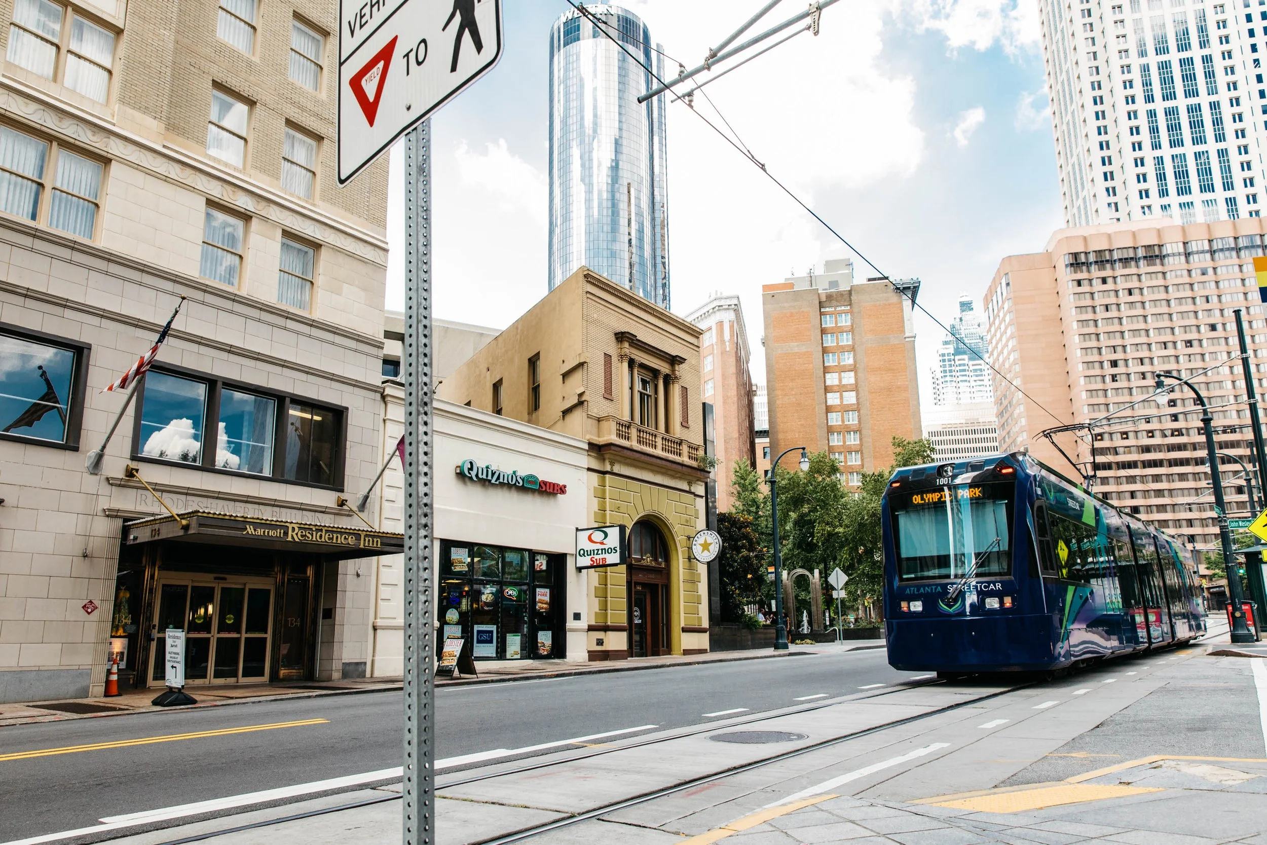A city street with tall buildings, a bus on the road, and a street sign indicating a pedestrian crossing and yield.