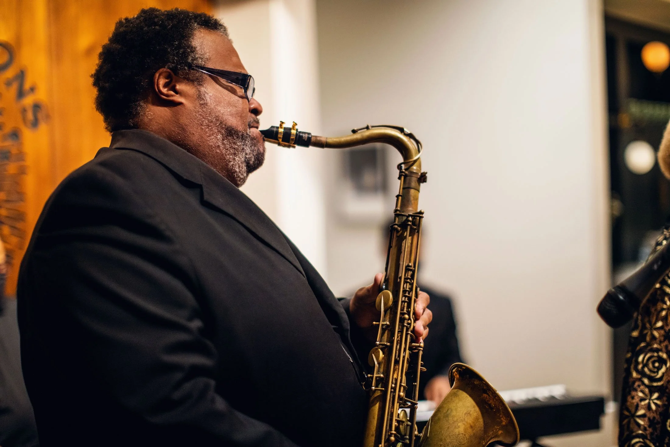 A man wearing glasses and a dark suit playing a saxophone, with a wooden wall and blurred background in an indoor setting.