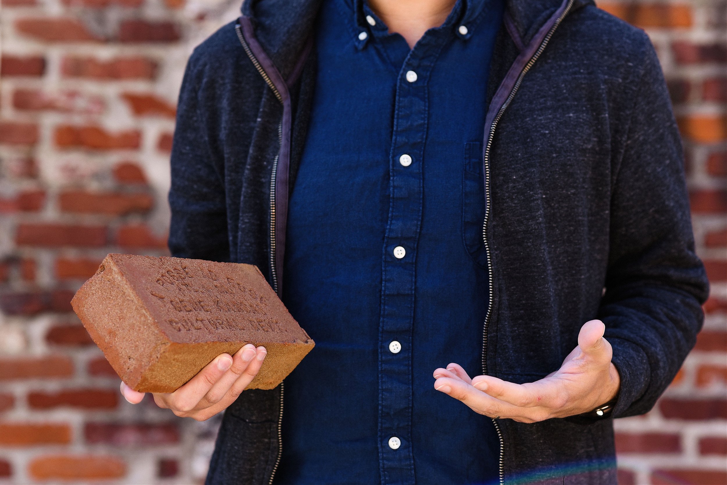 Person wearing a dark blue button-up shirt and dark hoodie, holding a brick.