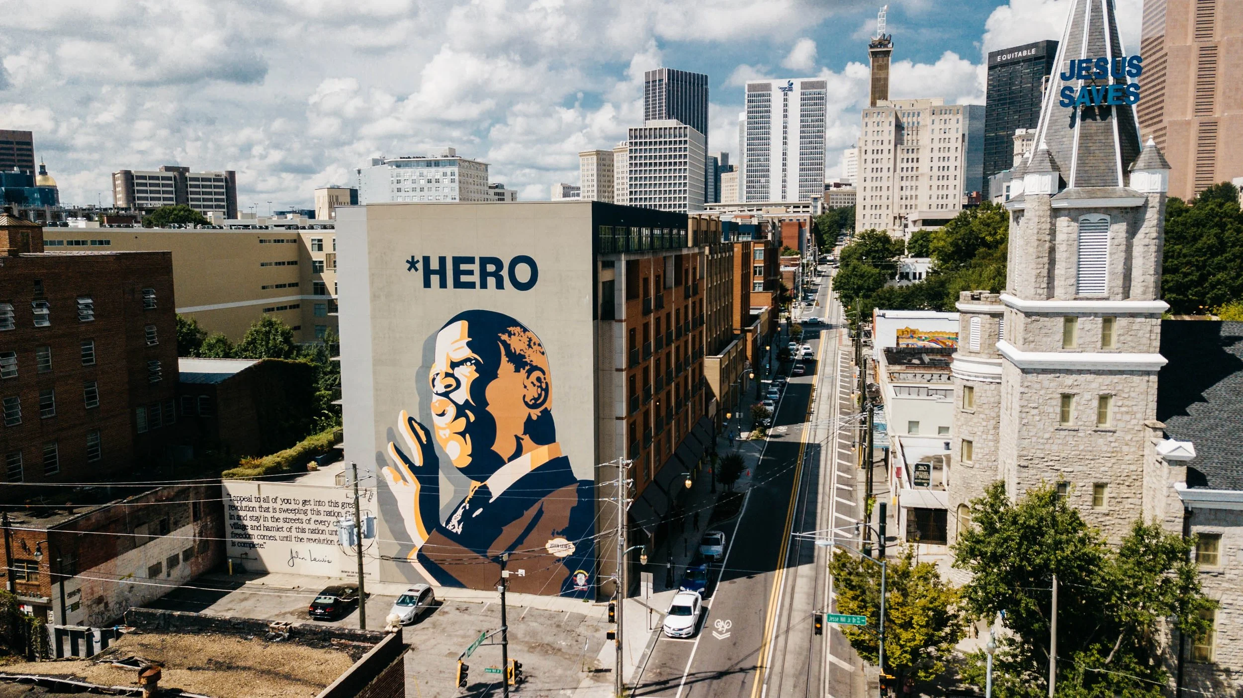 Cityscape view with tall buildings, including a prominent mural of a man with glasses and a hat, and a billboard that reads 'JESUS SAVES' against a cloudy sky.