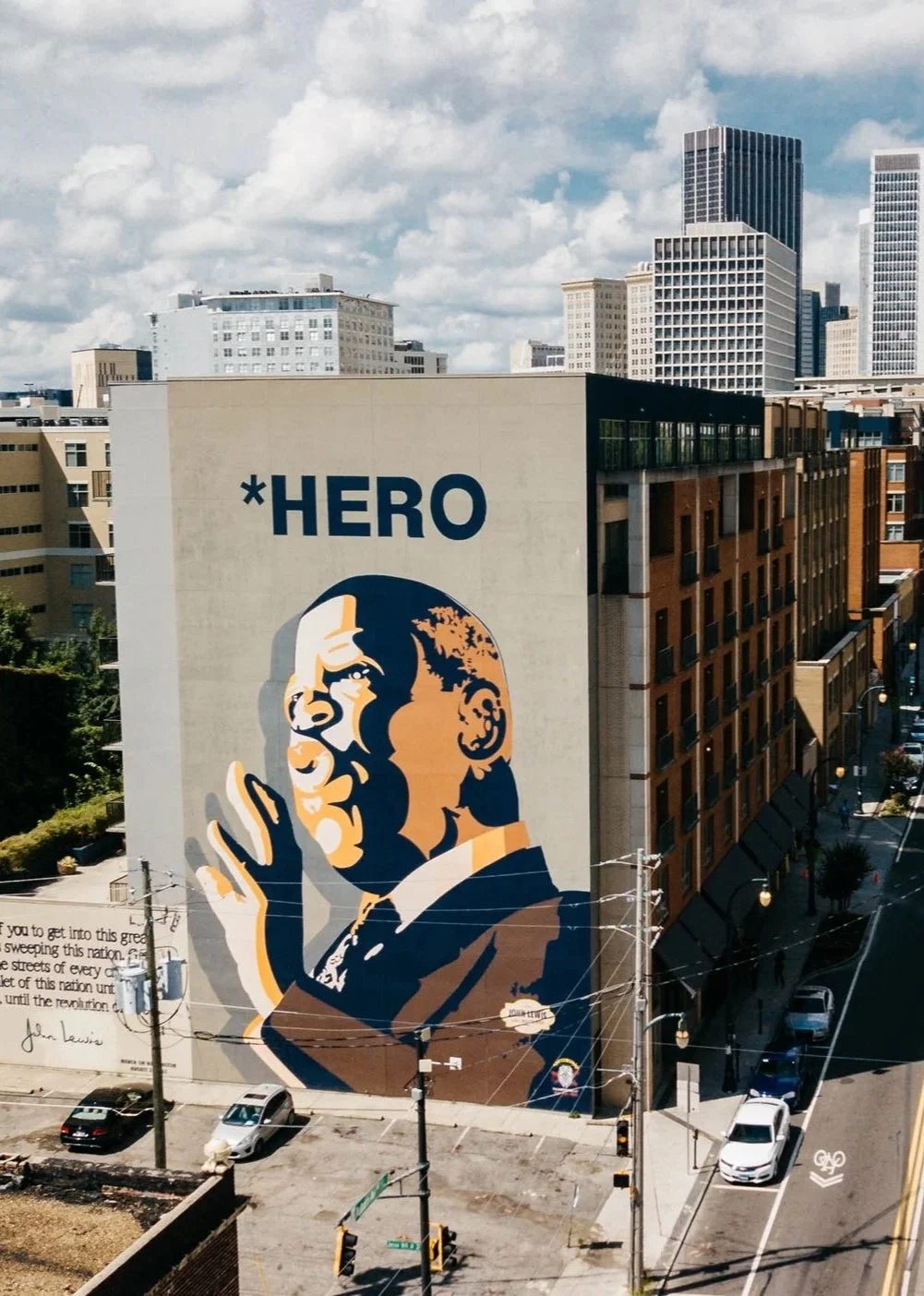 Large mural of a man in a uniform making a peace sign, with the word '*HERO' above it on a city building, with skyscrapers and streets below.