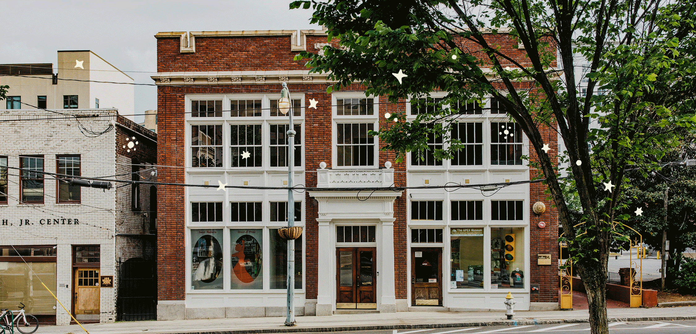 A historic 1910 multi-story brick building with large white-framed windows, a central entrance with wooden doors, and decorative elements, situated on a city street with a sidewalk, trees, and a streetlamp in front. Home to Constellations.