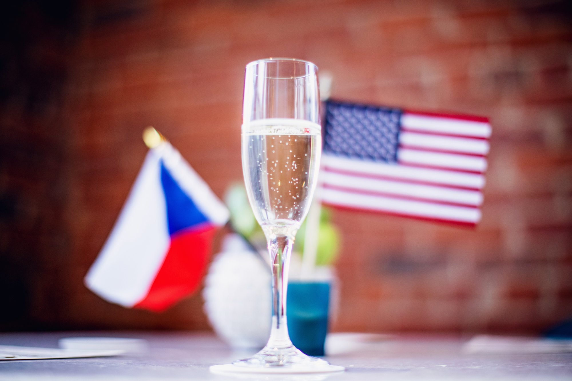 A champagne flute filled with sparkling water with bubbles, placed on a table. In the background, there are small flags of the United States and France, and a Greek flag, with a blurred brick wall behind them.