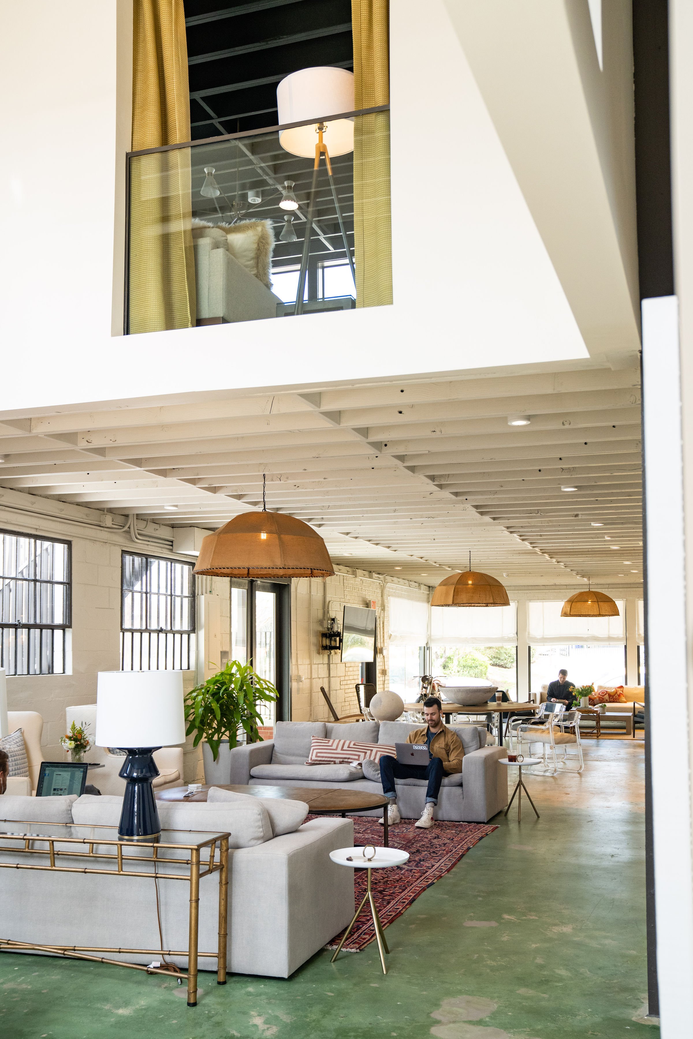 Open-concept industrial-style coworking space with high ceilings, large windows, modern furniture, and people working on laptops.