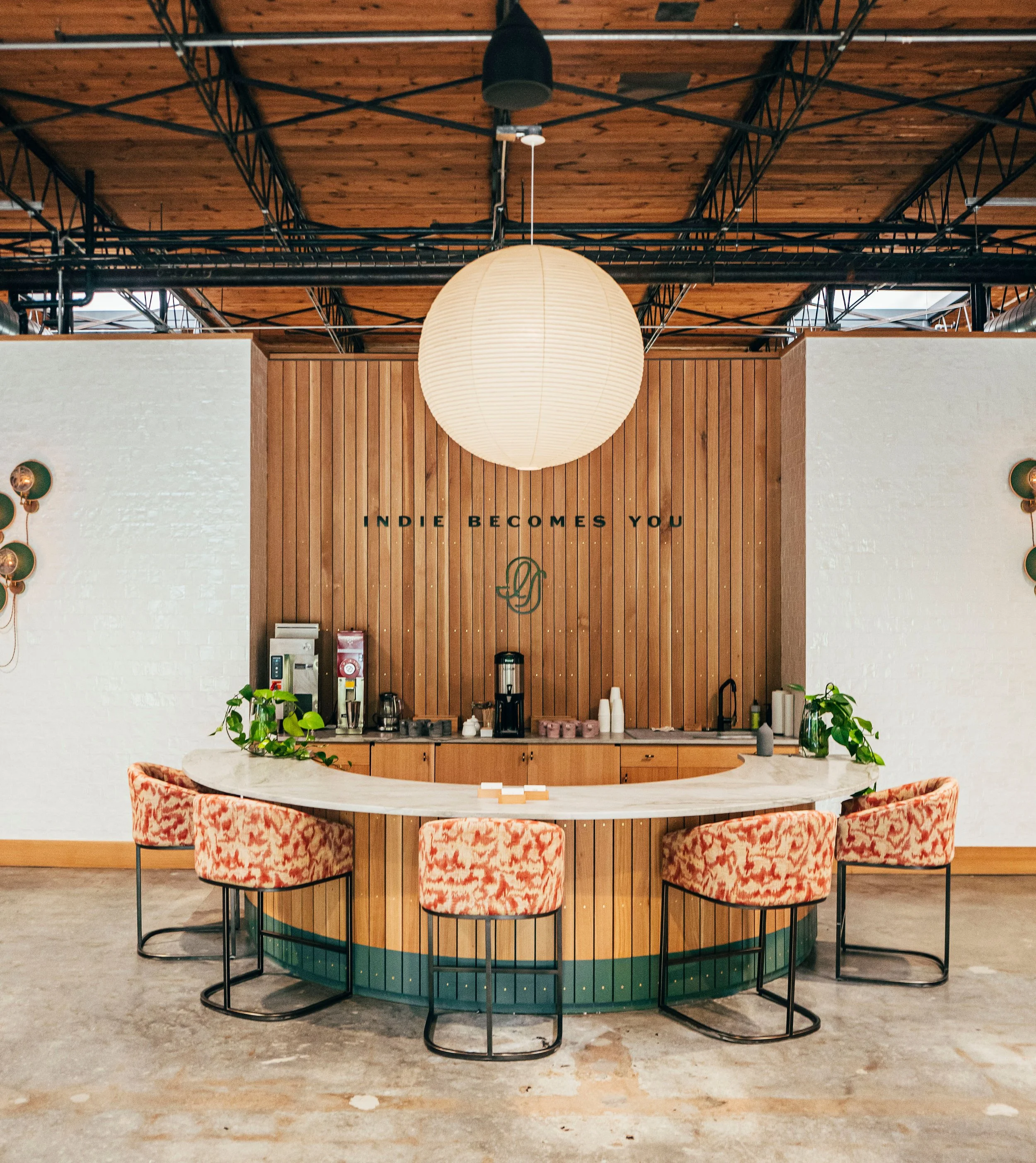 Interior view of a modern cafe with a rounded marble counter, decorative chairs with orange and white patterned upholstery, and a wooden wall with a round paper lantern hanging from a ceiling with visible black metal framework.