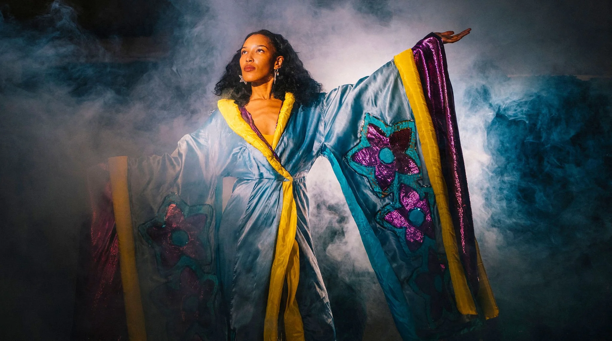 A woman in a colorful, flowy costume with butterfly patterns, standing with arms extended, surrounded by mist or smoke and dramatic lighting.
