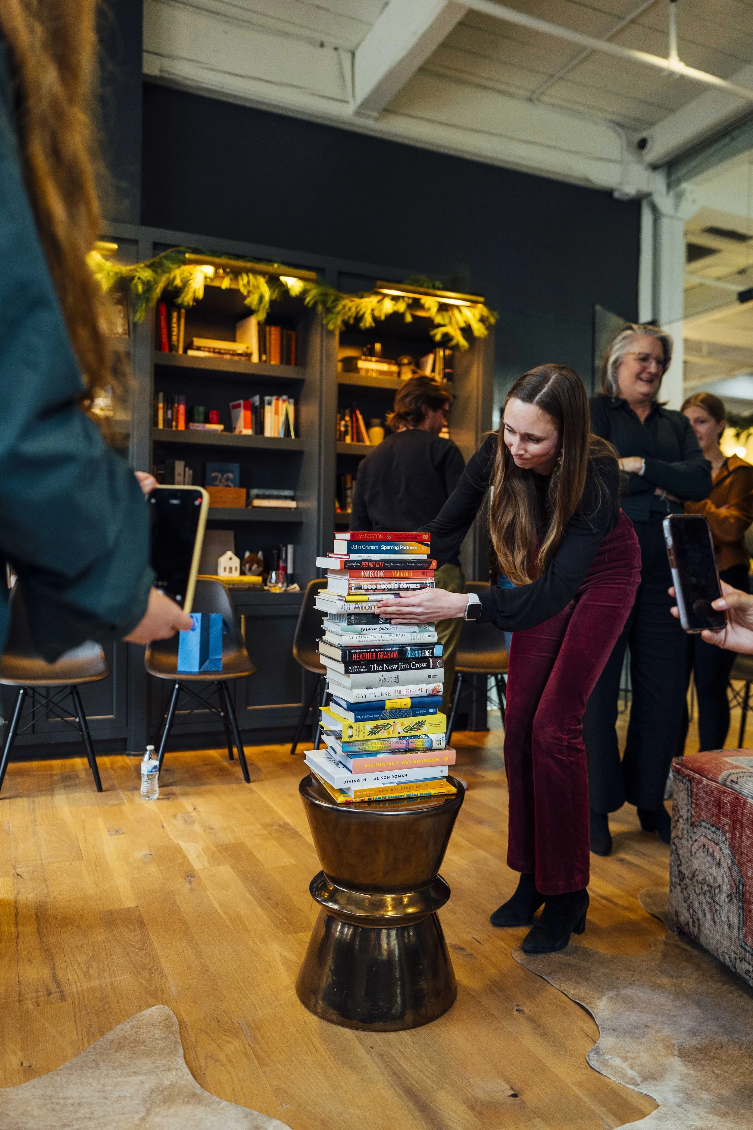 A woman with long brown hair arranging a stack of books on a round table in a cozy, warmly lit room with bookshelves and other people in the background.