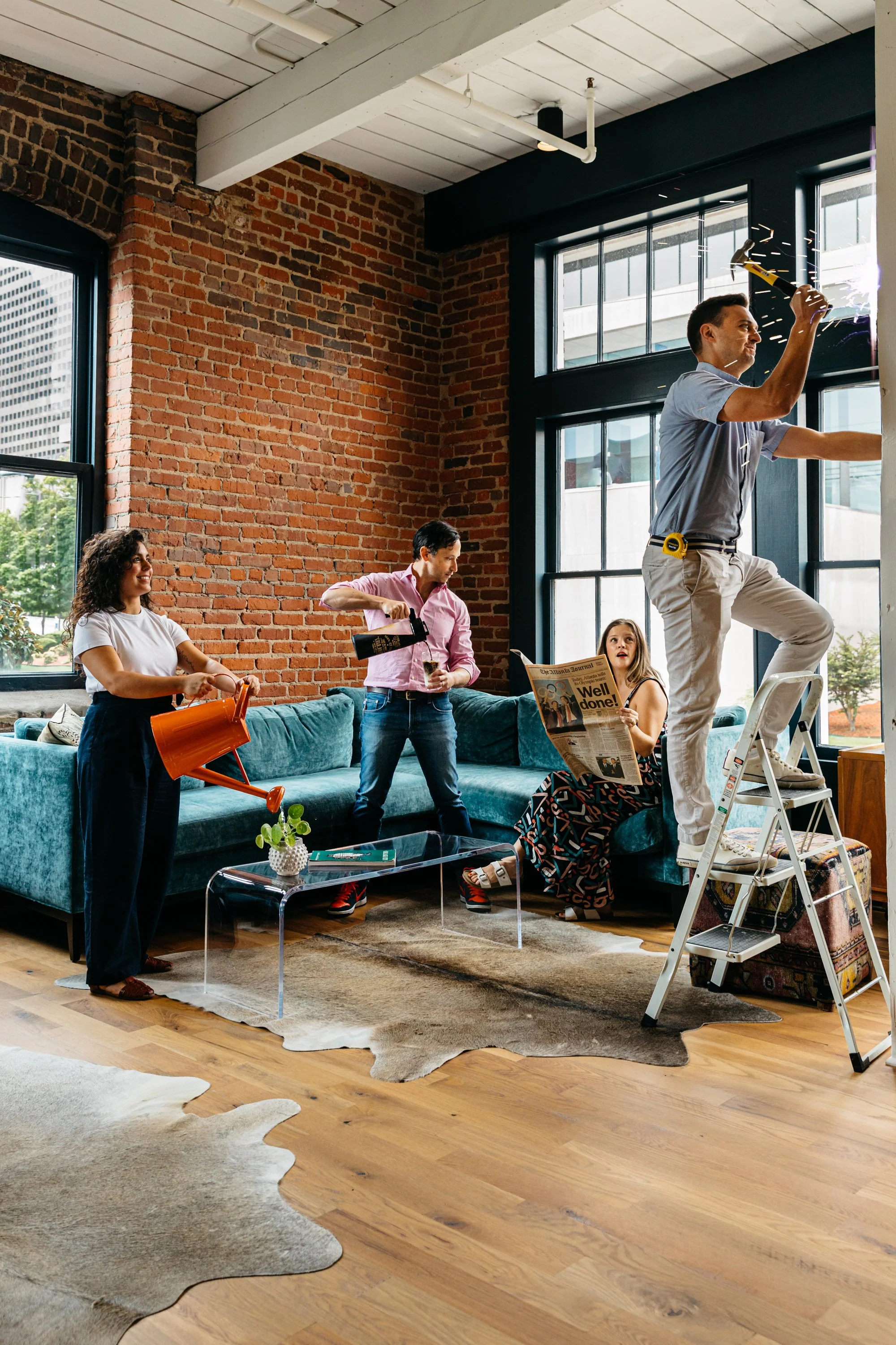 A group of four people in a loft-style living room with exposed brick walls and large windows. One person is on a ladder using a tool, others are watching or chatting. The room has a sofa, coffee table, and cowhide rugs.