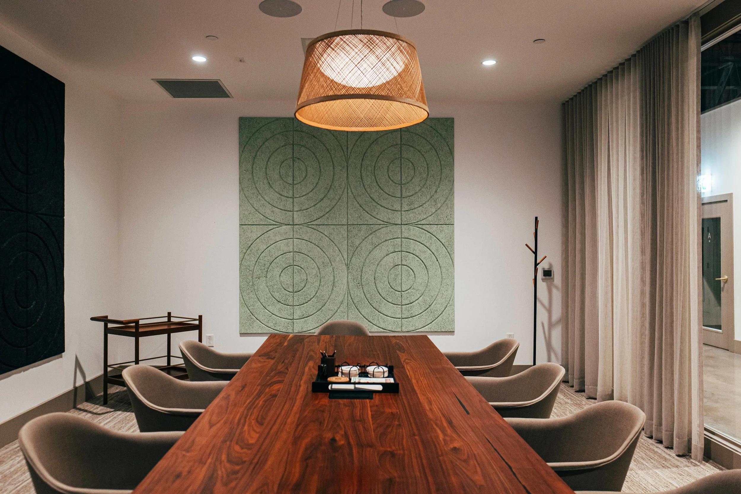 A modern conference room with a wooden table, beige chairs, a large woven pendant light, green artwork on the wall, and curtains covering windows.