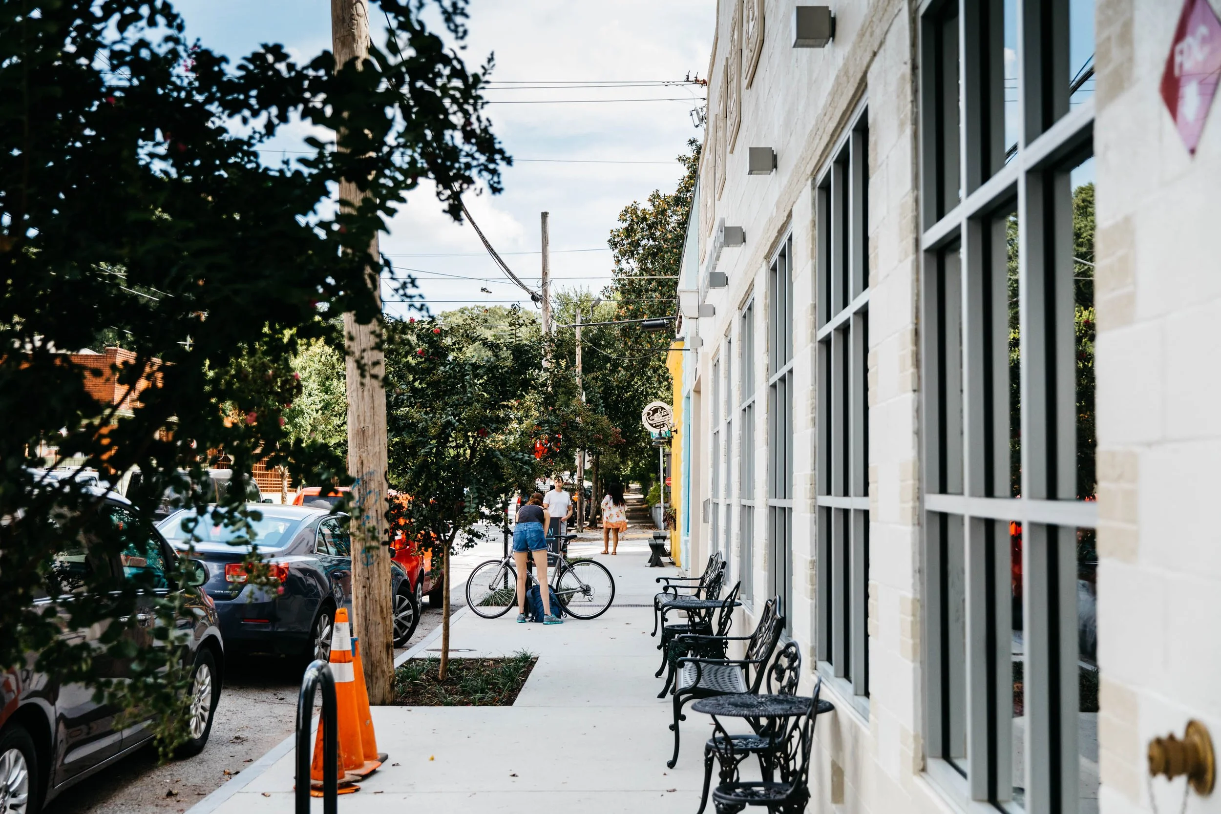 Sidewalk cafe with empty black metal chairs outside a beige brick building, people walking on the street, parked cars, trees, and utility poles in the background.