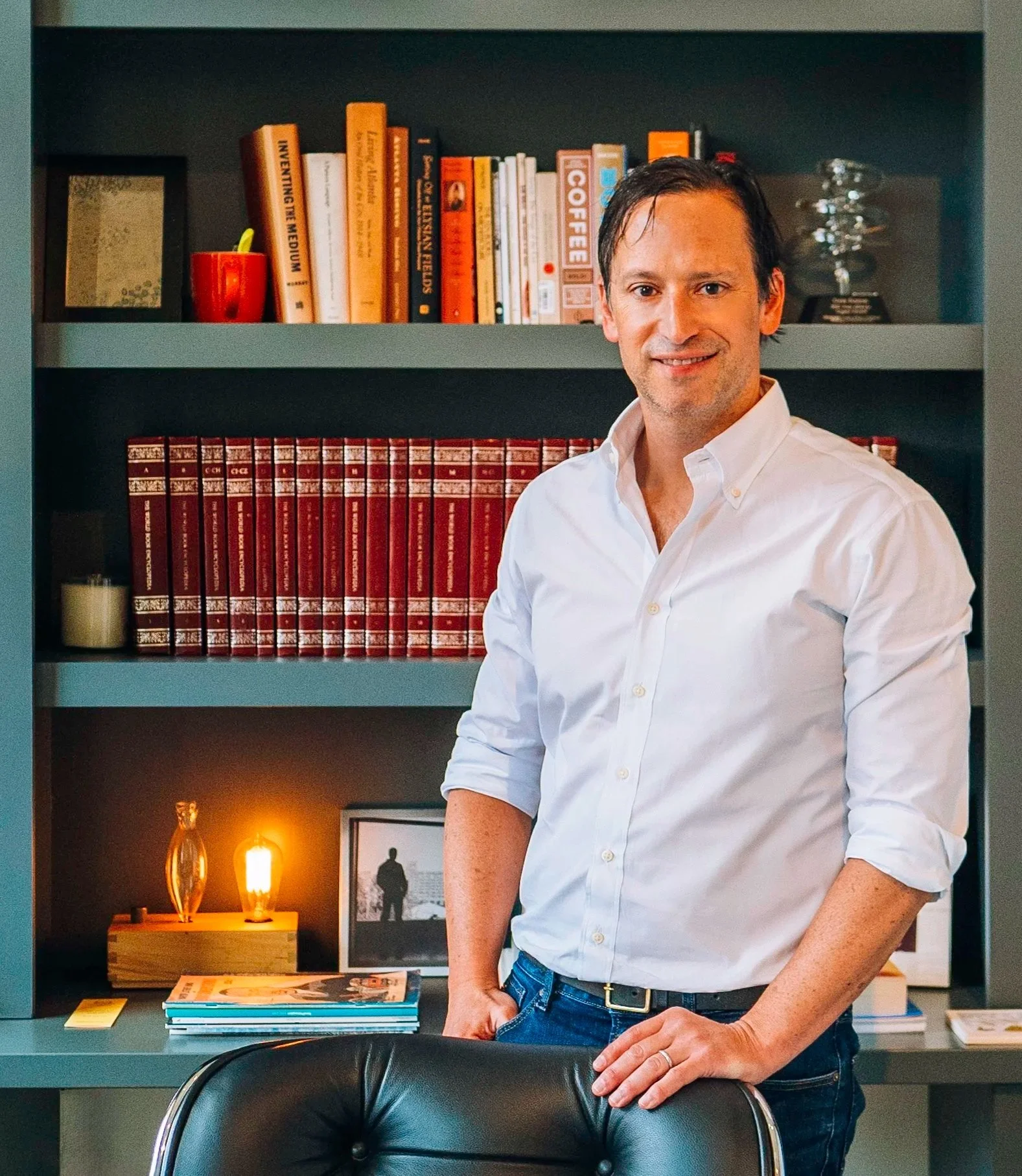 A man wearing a white button-down shirt, standing in front of a bookshelf with books and decor, inside a room with warm lighting.