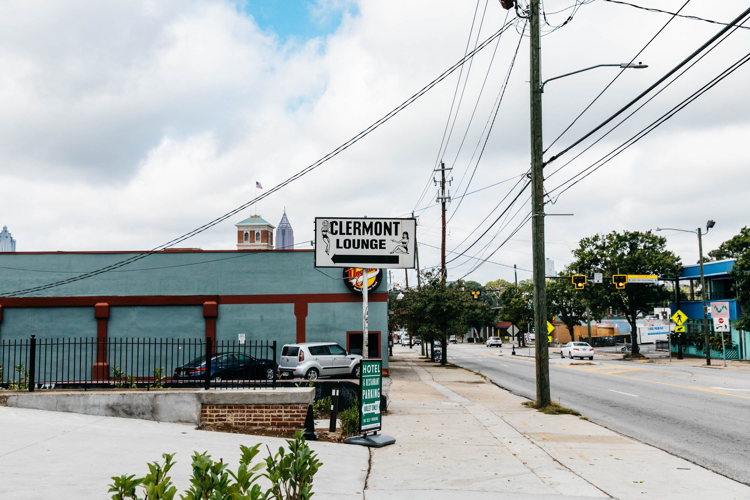 A street scene with a motel, parking, and hotel sign. Utility poles and wires are overhead. In the background, the city skyline is visible under a cloudy sky.