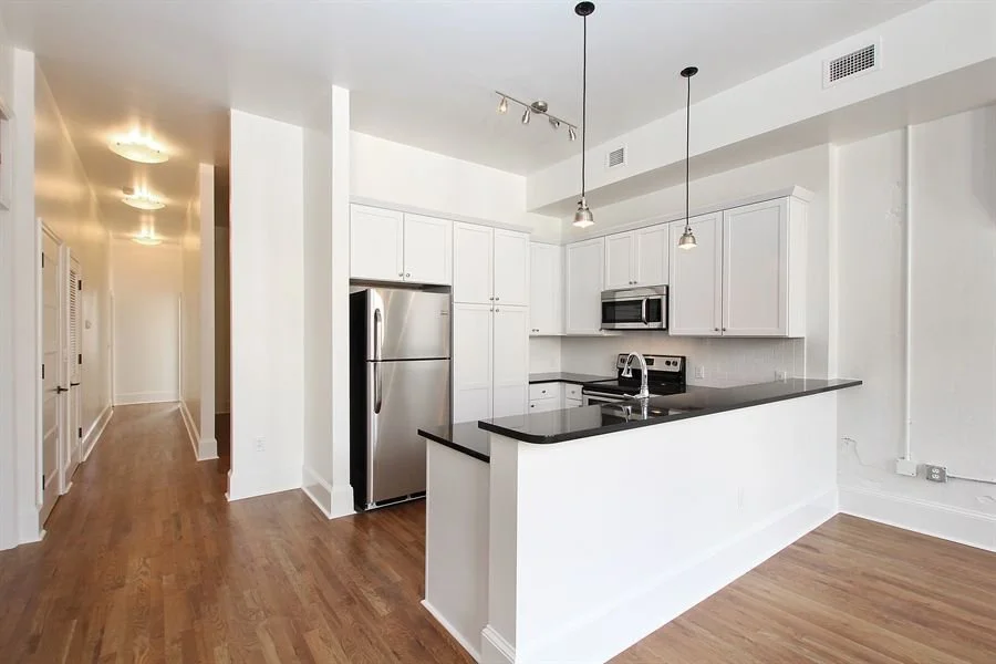 Modern kitchen with white cabinets, stainless steel refrigerator and microwave, black countertop, and wooden floors. Two pendant lights hang over the counter, and a hallway with ceiling lights is visible on the left.