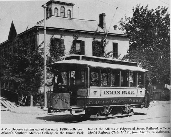 Black and white photo of an early 1890s electric streetcar labeled 'Inman Park' on Route 7, with a man standing on the street next to it, passing a large historic brick building with multiple stories, arched windows, and a tower-like structure on the roof, surrounded by trees.