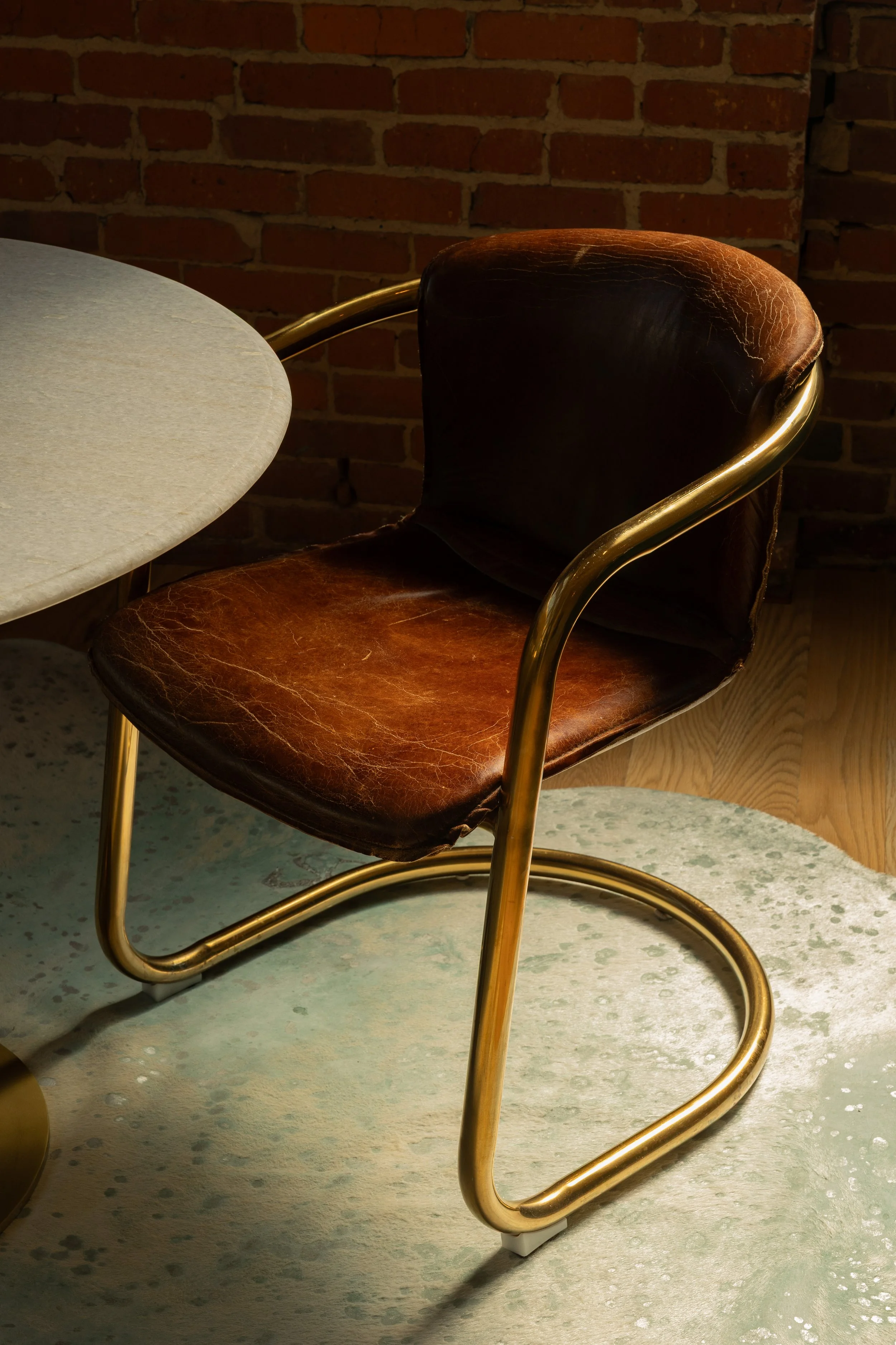A corner of a round marble table with a smooth, light gray surface. Next to it is a vintage brown leather chair with visible cracks, featuring a curved gold metal armrest and a gold metal frame. The background has a red brick wall.