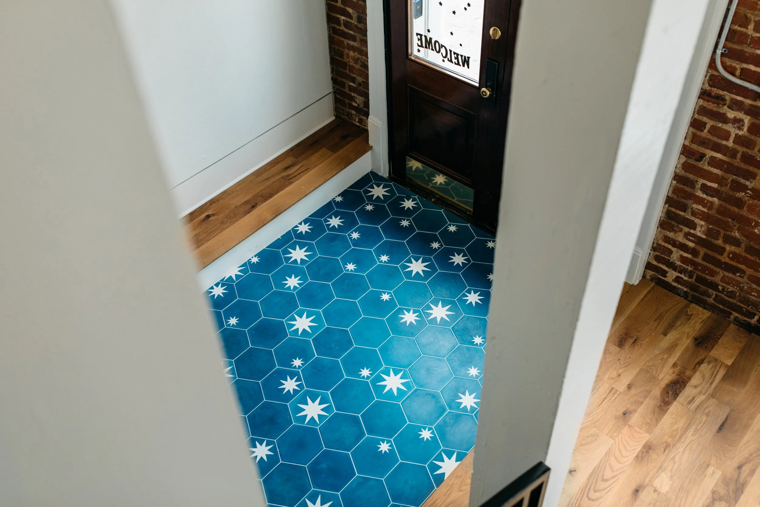An entryway with a dark wooden door featuring a glass panel with the word 'Welcome' and small star decals. The floor has blue tiles with white star patterns, and there is a brick wall on one side and a white wall on the other. A wooden step is presen