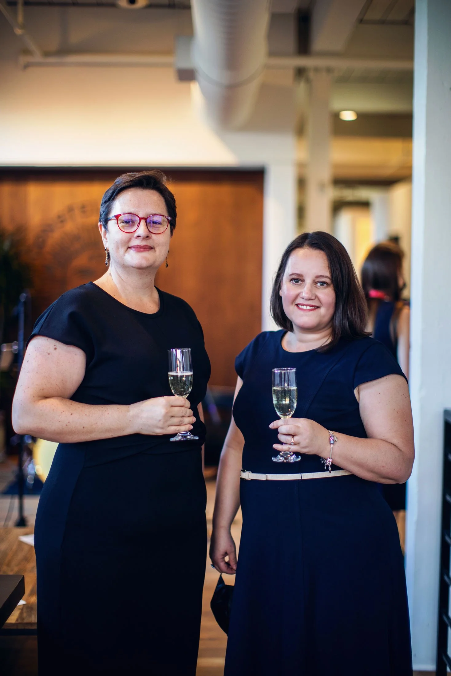 Two women in dark dresses holding champagne glasses at an indoor social event. One woman has short dark hair and wears red glasses, the other has shoulder-length dark hair. The background is blurred with warm lighting and other people visible.