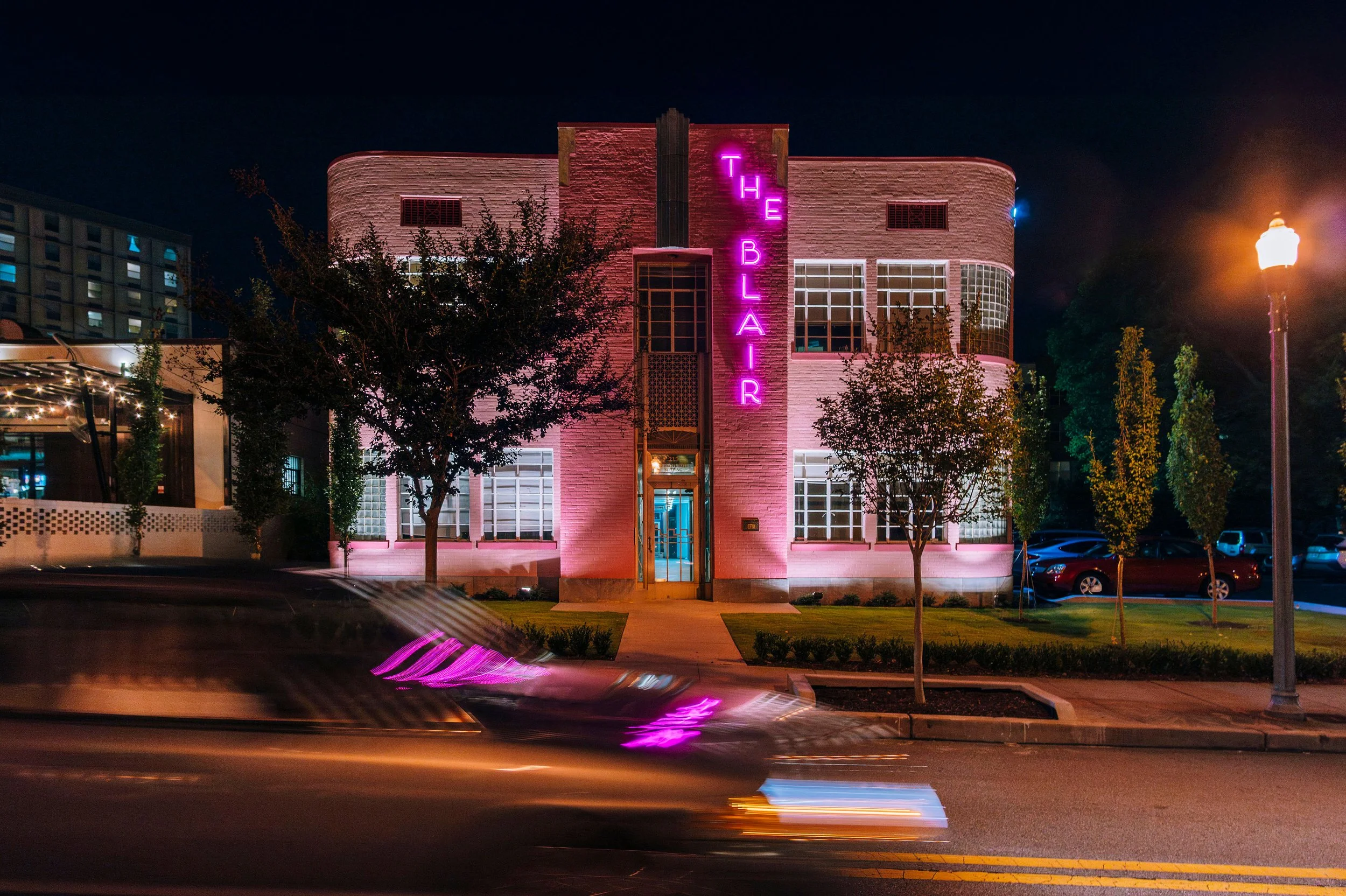 Night view of a building with pink neon sign reading 'THE BLAIR' and illuminated windows. A streetlamp and some trees are in the foreground, and a passing car creates light streaks in the image.
