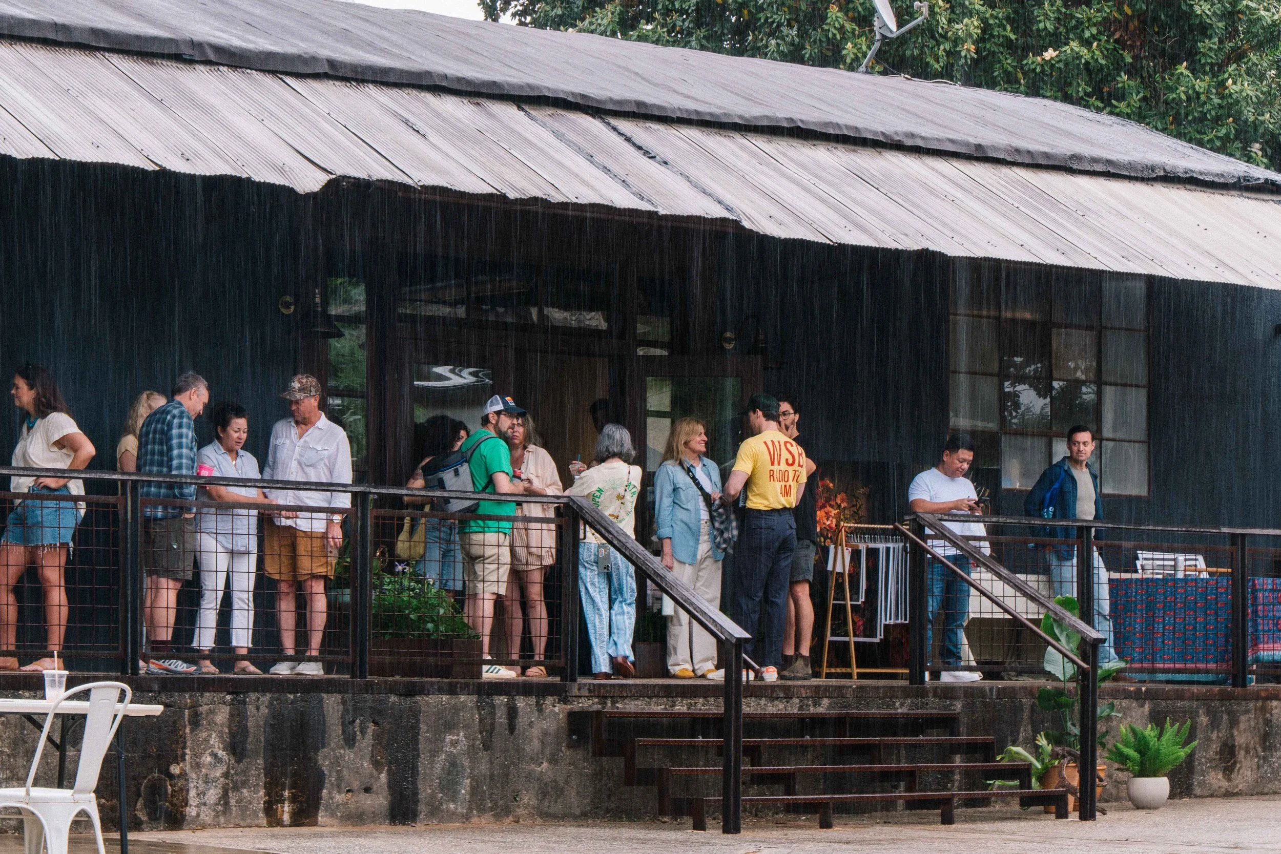 A group of people gathered outdoors on a balcony or deck, engaging in conversations, with some looking at their phones, under a roof with rain falling.