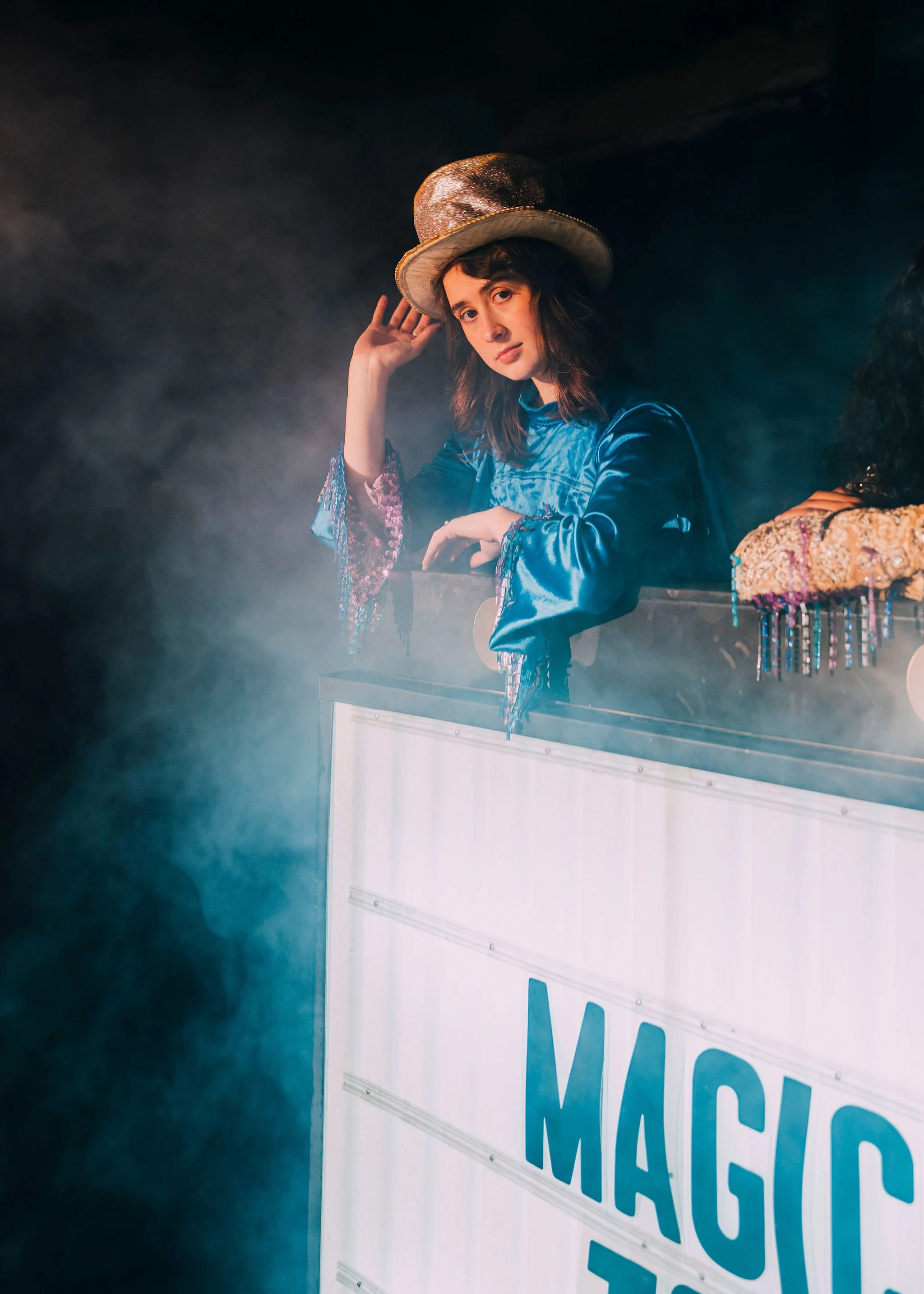 Person wearing a shiny, large, golden hat and a shiny blue dress, posing next to a white barrier with blue text, in a smoky environment.