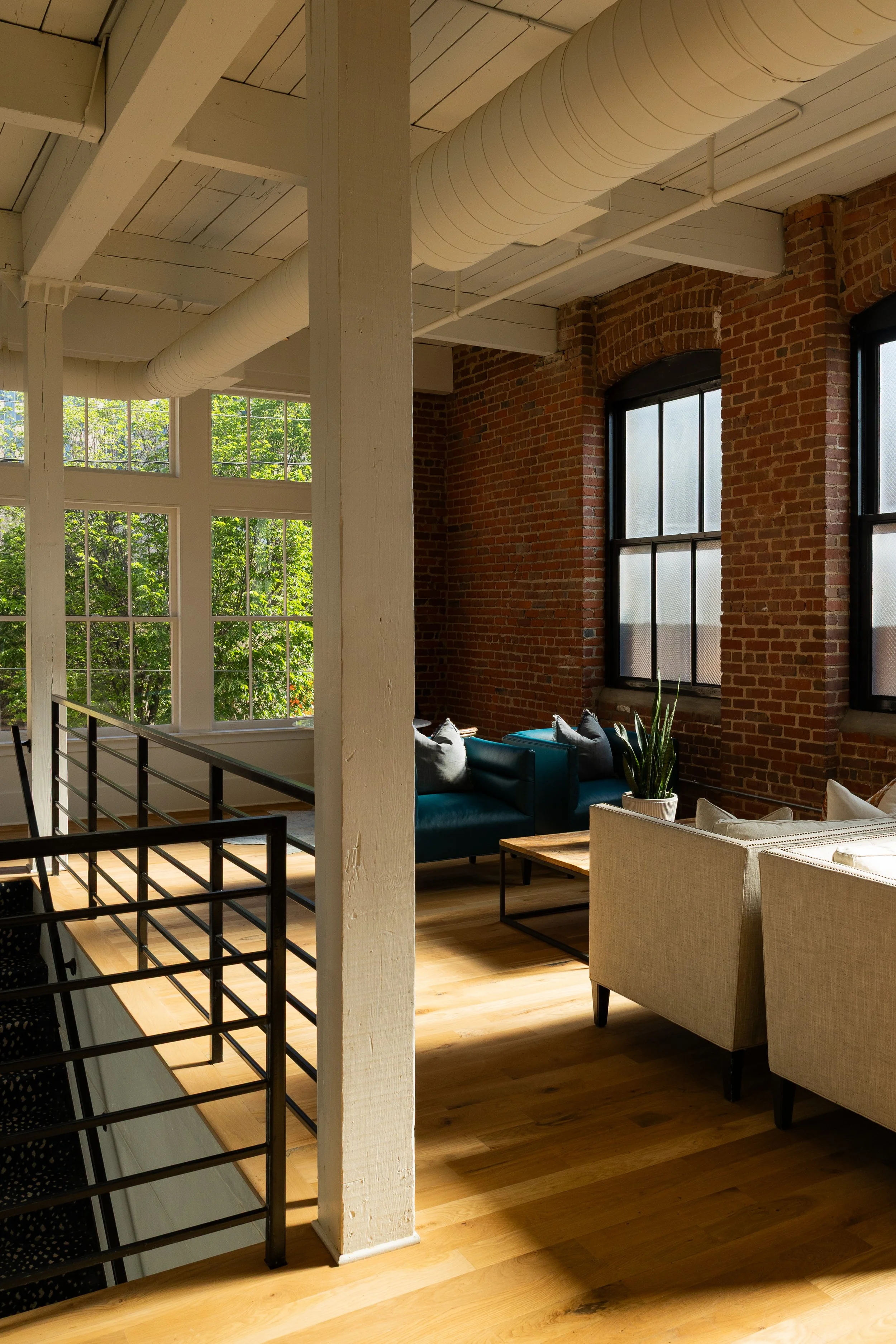 Bright living room with exposed brick walls, large windows with a view of greenery, and a mix of modern and vintage furniture including a beige armchair, blue sofa, and potted plant on a wooden table.