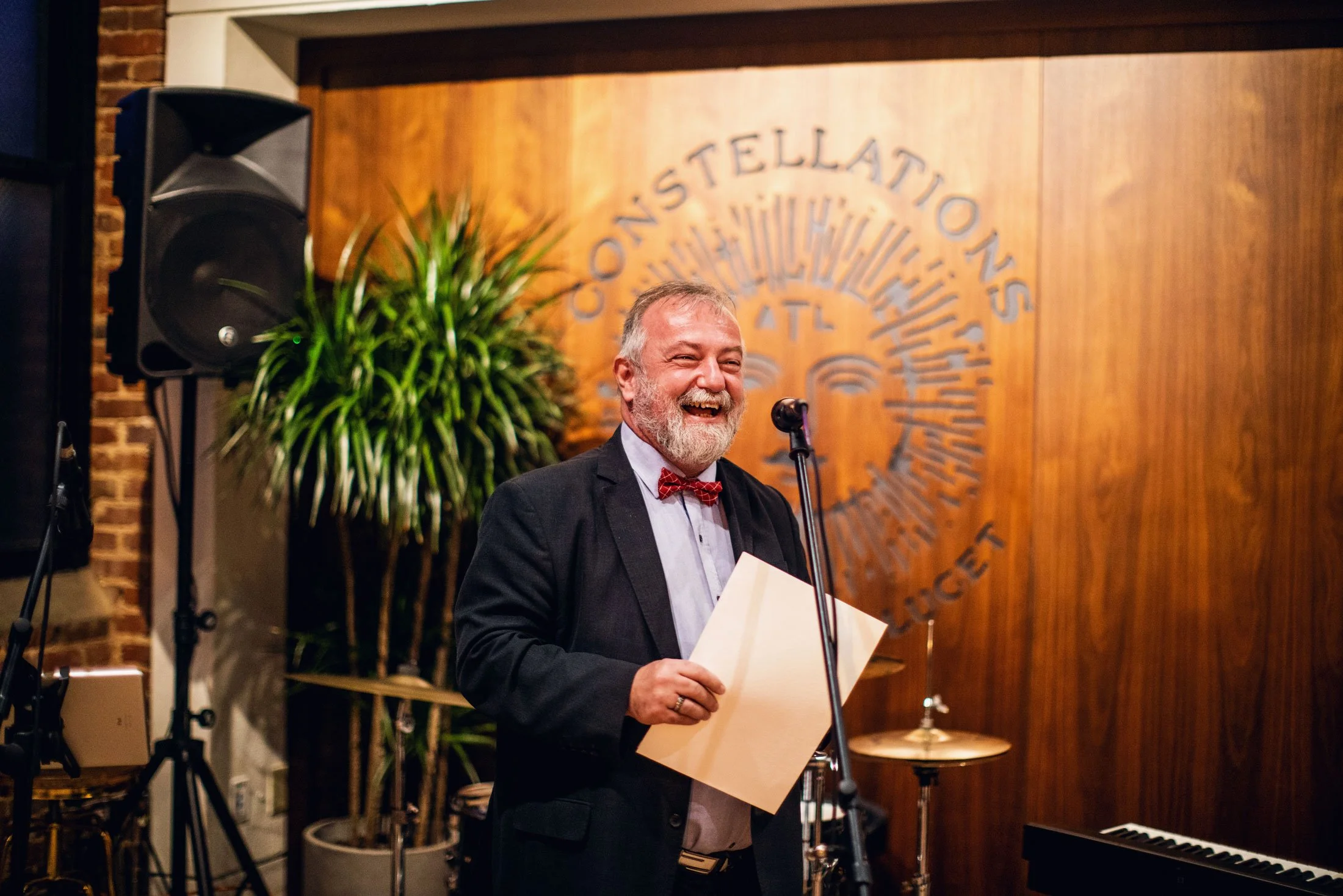 A man with gray hair and a beard, dressed in a suit and bow tie, smiling and holding papers on a stage with a musical instrument and a large wooden logo in the background.