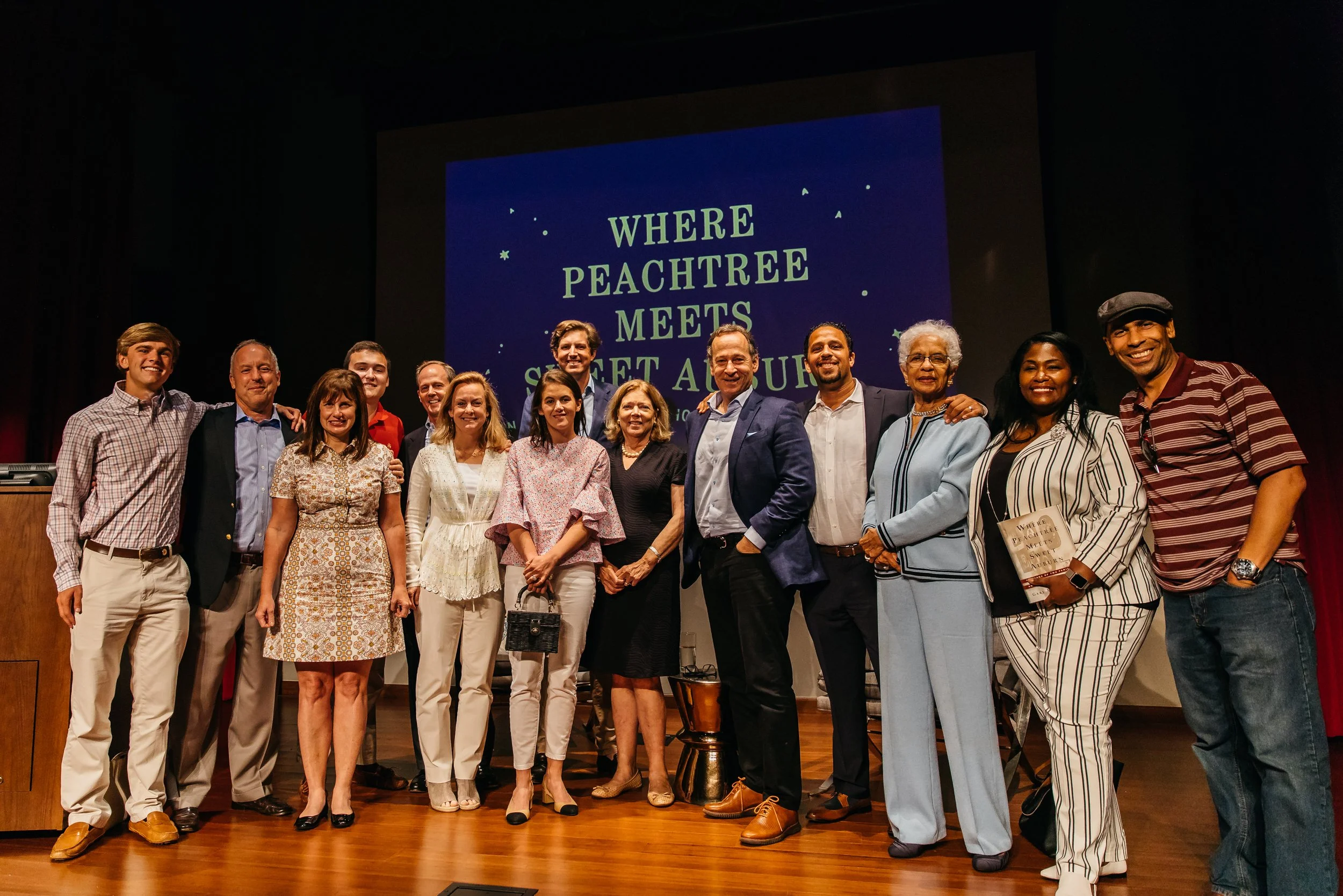 Group of people standing together on stage, smiling, with a large screen behind them displaying the title 'Where Peachtree Meets Sweet Auburn'.
