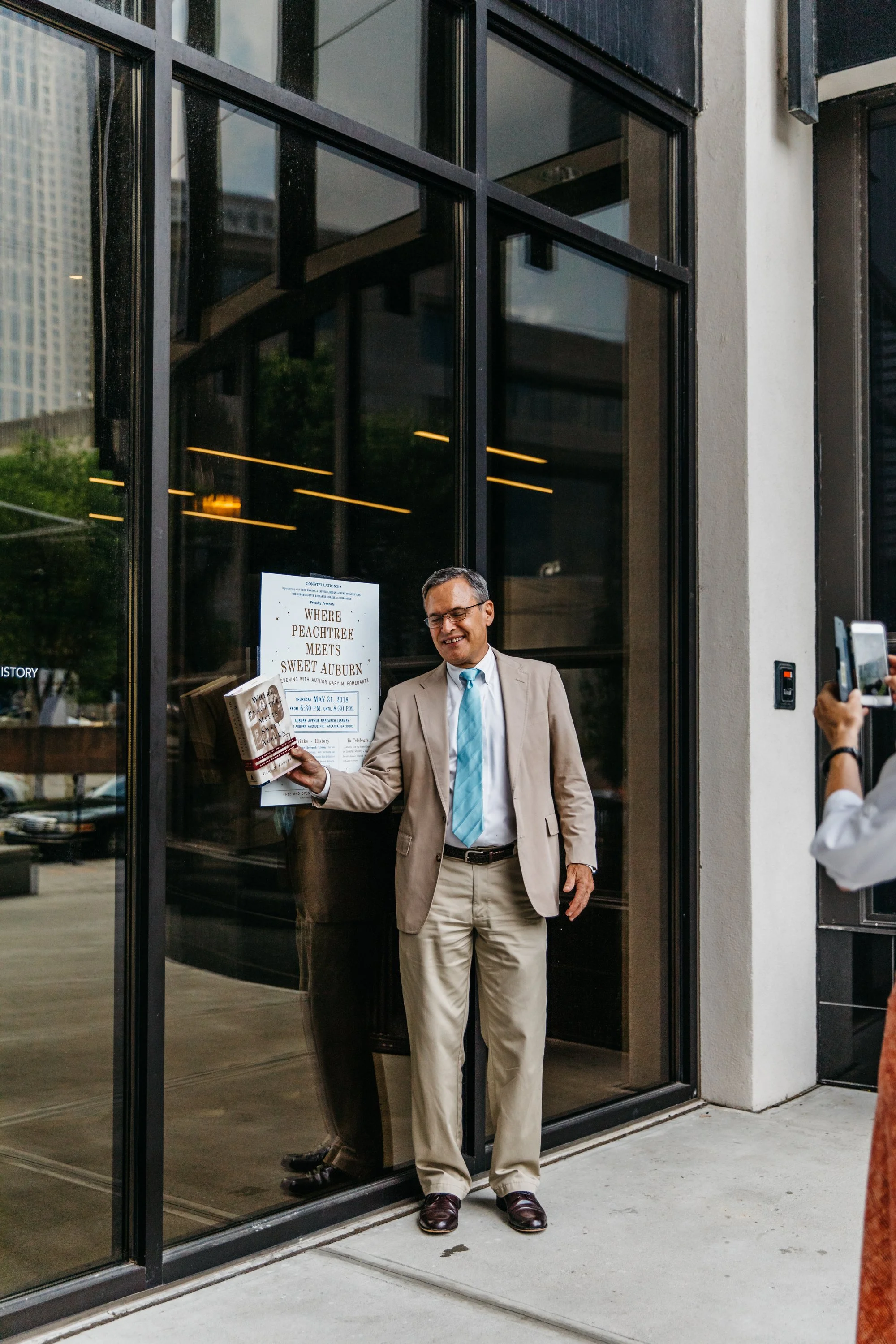 A man in a beige suit with a light blue tie and dark brown shoes smiling as he walks out of a glass building, holding a book. A person on the right is taking a photo or video of him.