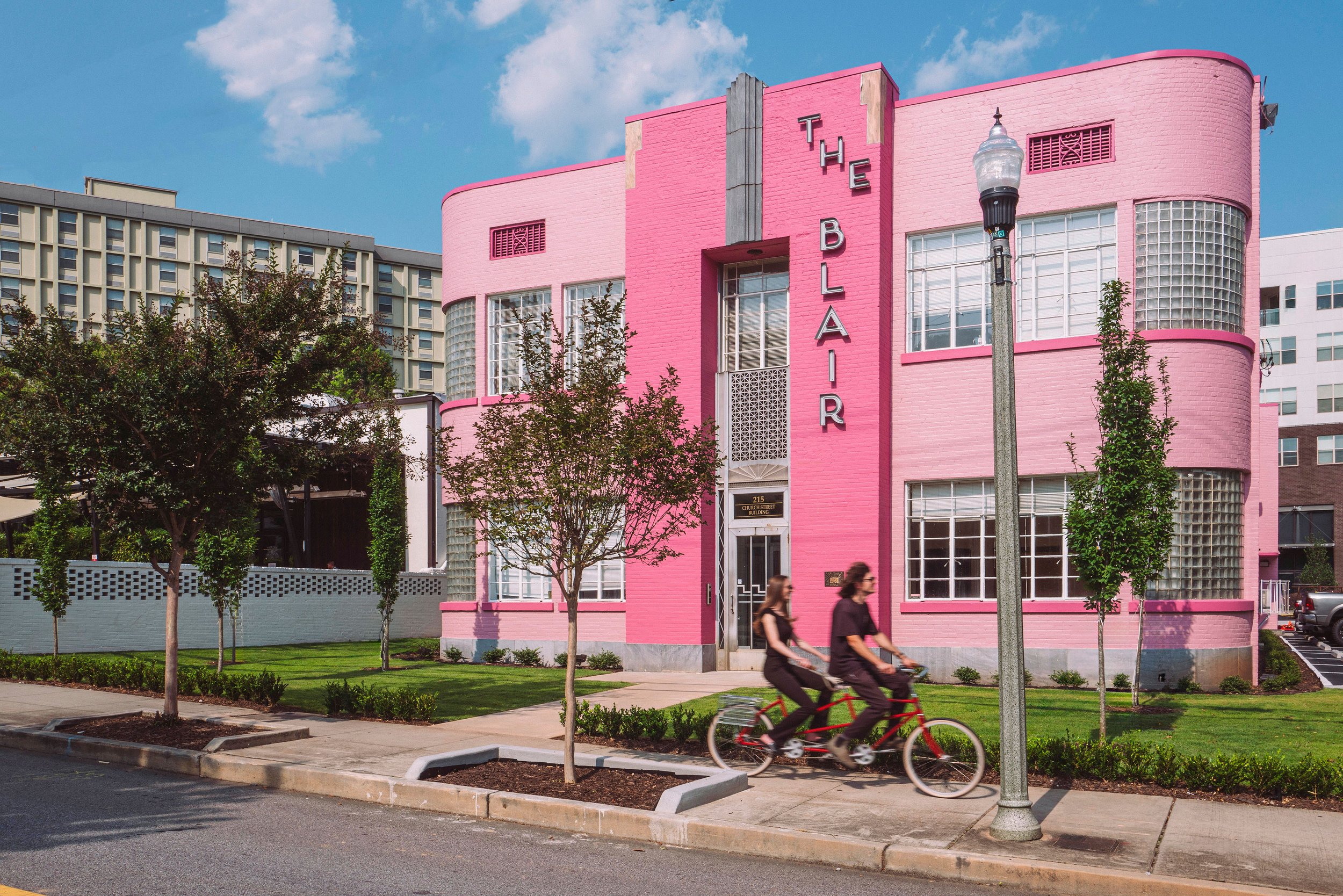 A pink Art Deco style building with the sign "The Blar" on the front, trees and people riding bikes in the foreground, and a blue sky with clouds in the background.