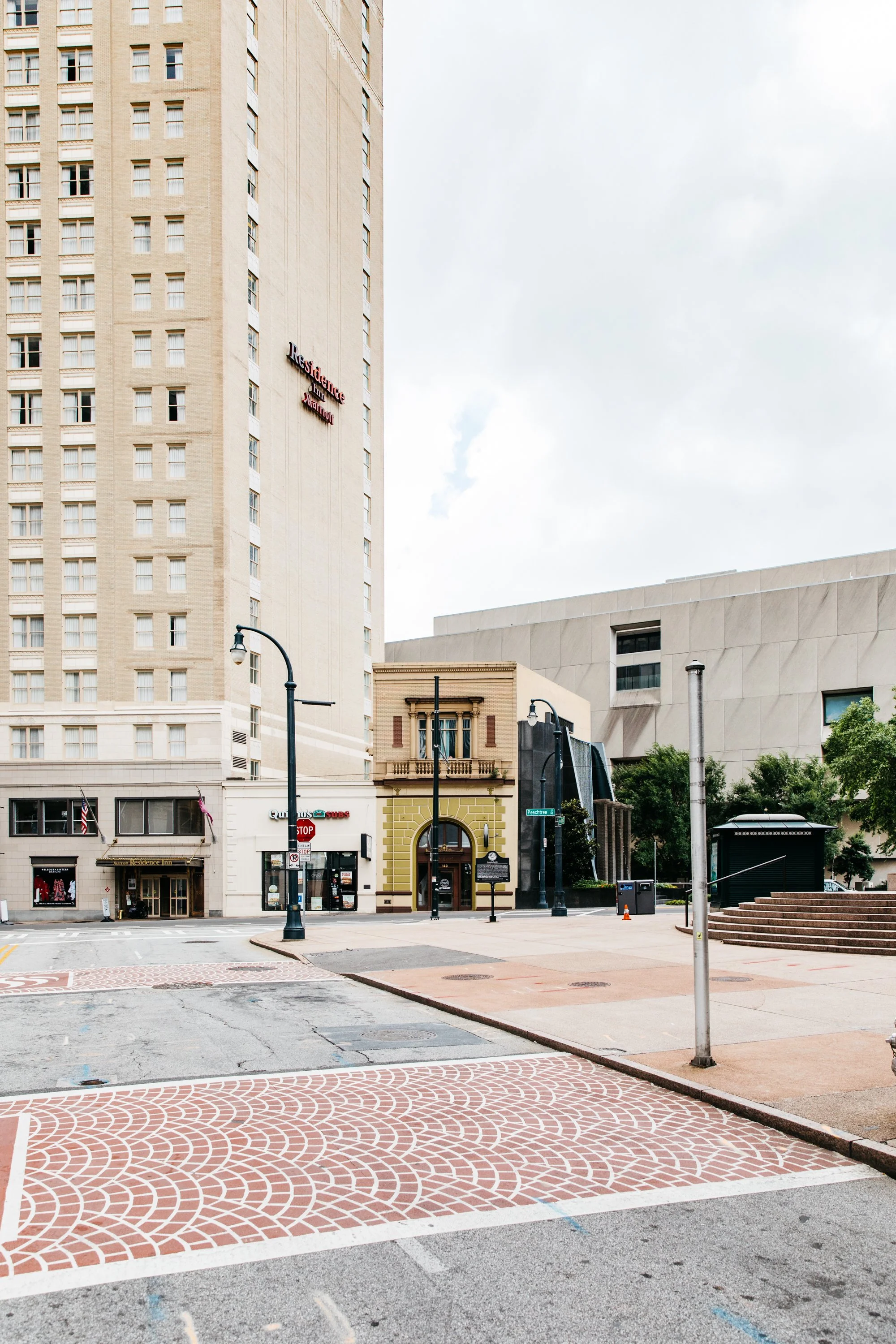 An urban street scene with tall buildings, storefronts, street lamps, and a patterned crosswalk on a cloudy day.