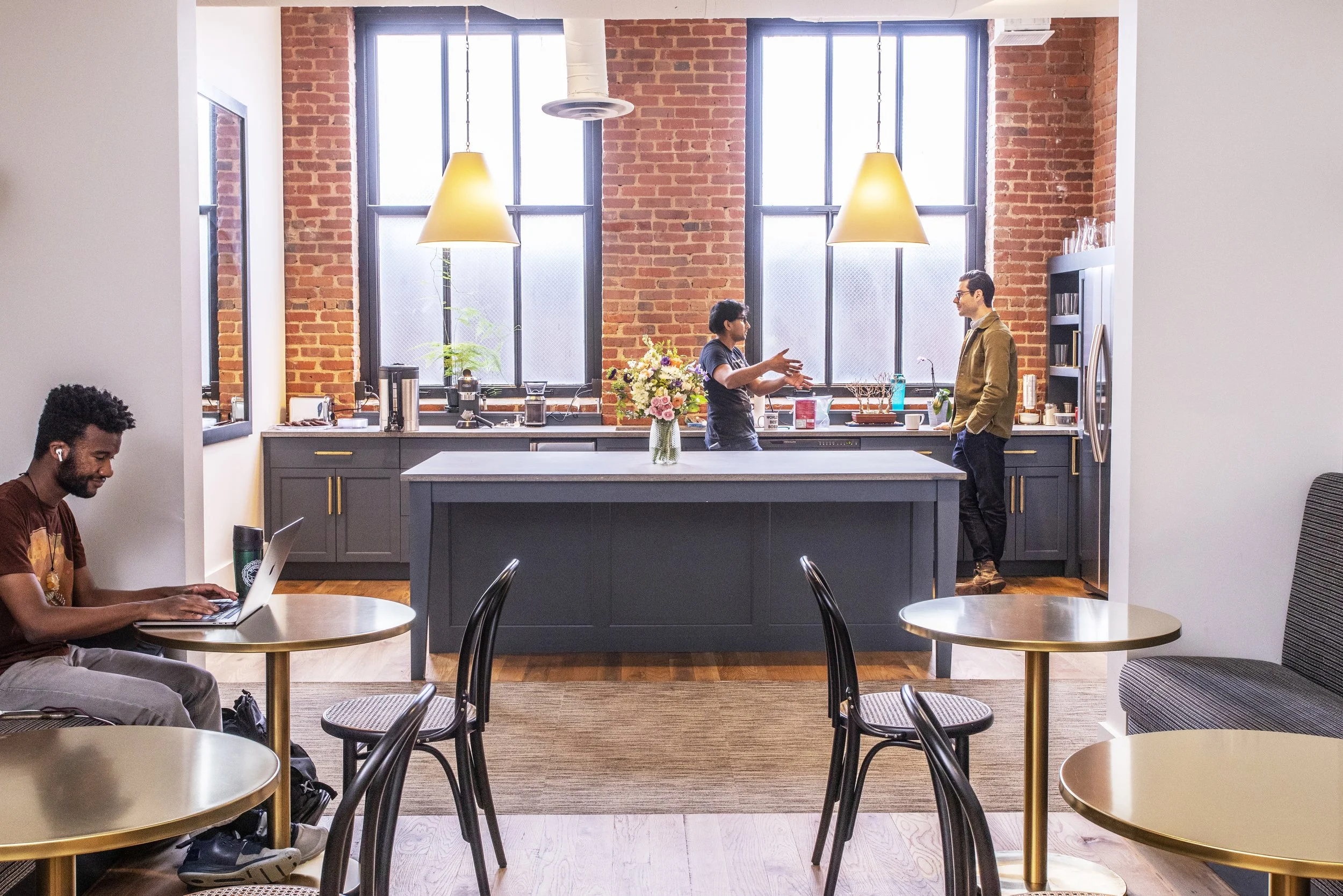 People in a kitchen and eating area of a modern apartment with exposed brick walls, large windows, and hanging pendant lights. One person is working on a laptop at a round table, while two others are standing and talking by the kitchen counter.