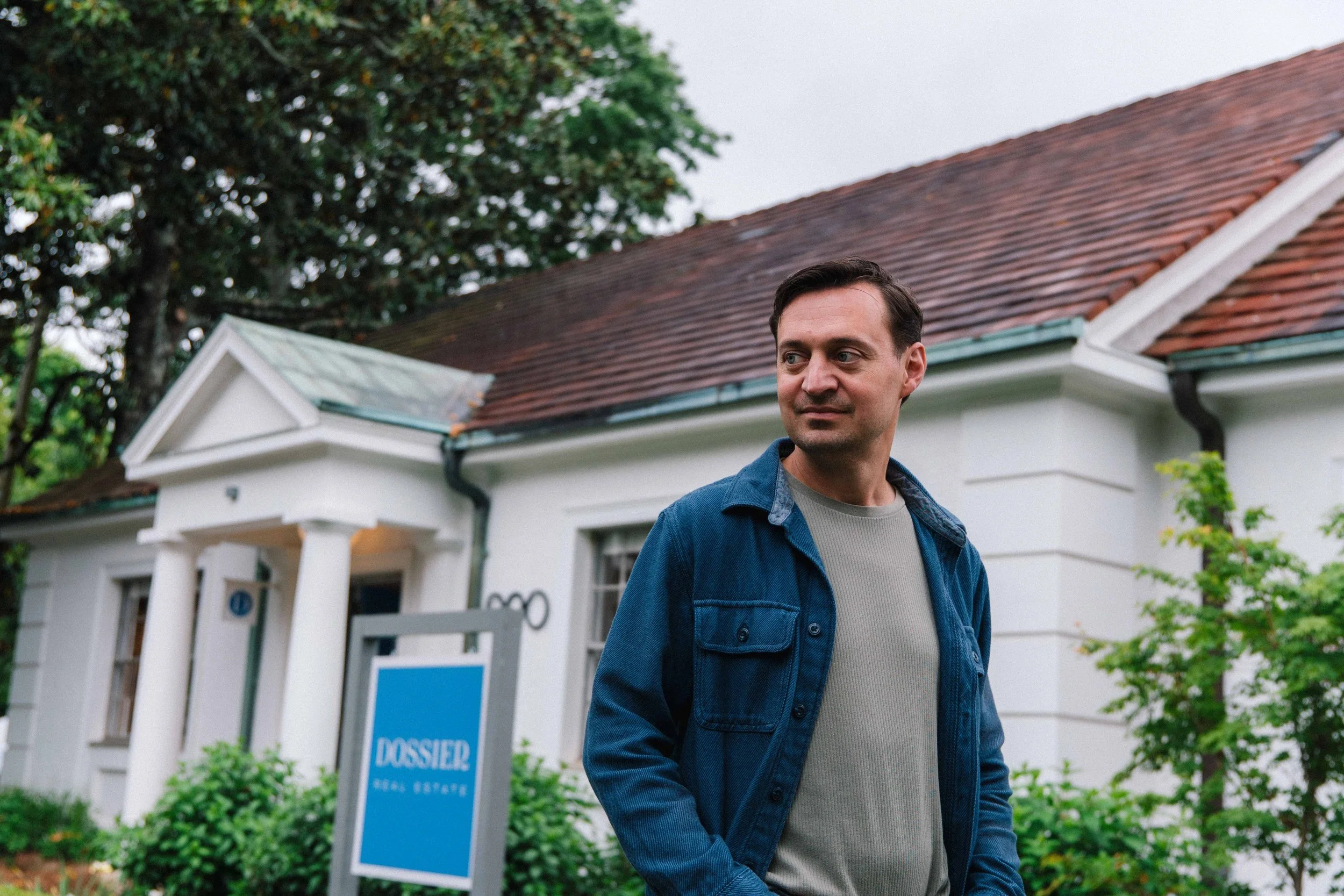 A man in a blue jacket and gray shirt walking past a real estate sign in front of a white house with a red tile roof and green trees in the background.