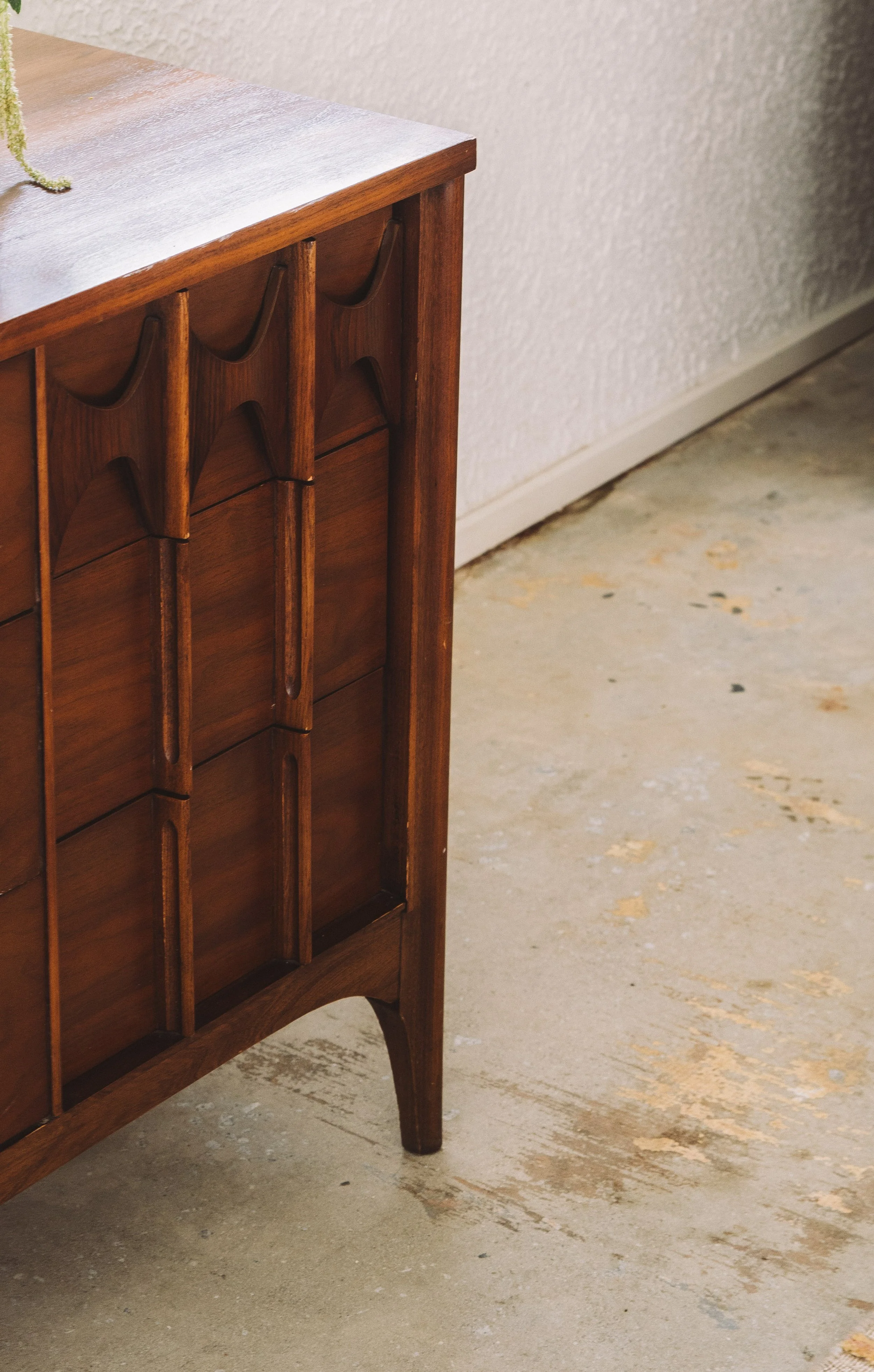 Part of a wooden piece of furniture with a scalloped design on its side, standing on four curved legs, against a light textured wall and a concrete floor.