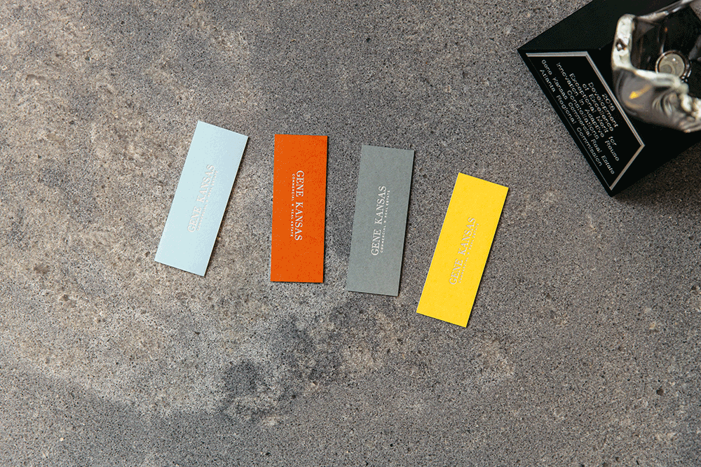 Four small rectangular sample cards in white, red, gray, and yellow, each with the text 'Gene Kansas' printed on them, are arranged on a textured gray surface next to a black box.