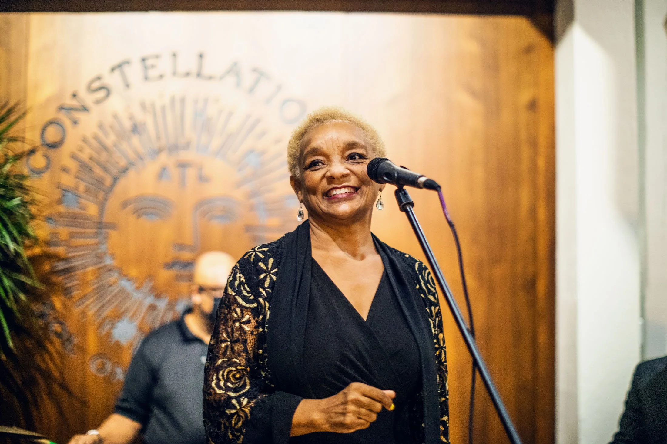 Smiling woman with short, blonde hair standing at a microphone in front of a wooden wall with partially visible text in the background.