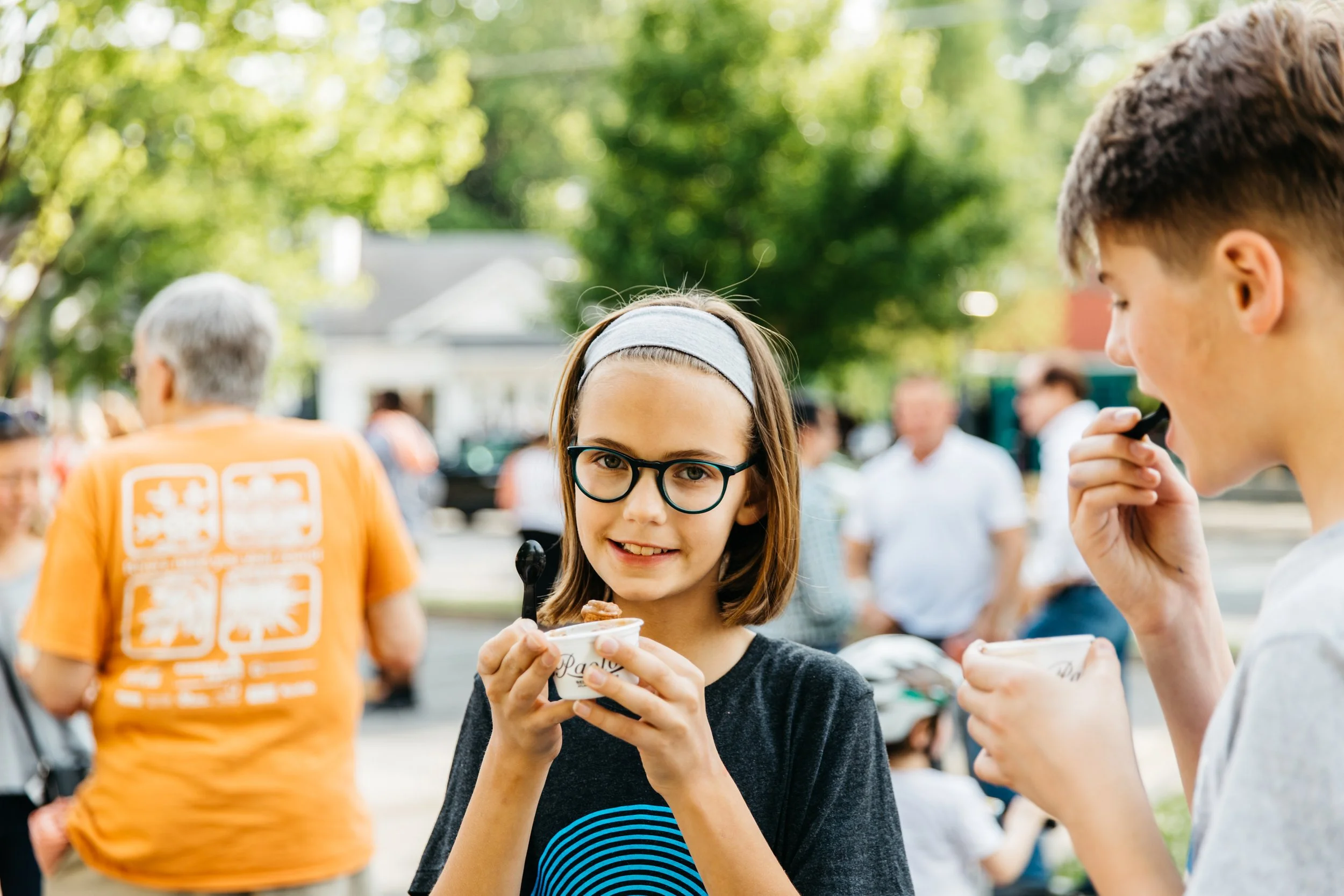 Young girl with glasses and dark hair wearing a gray headband enjoying ice cream at an outdoor event with people and trees in the background.