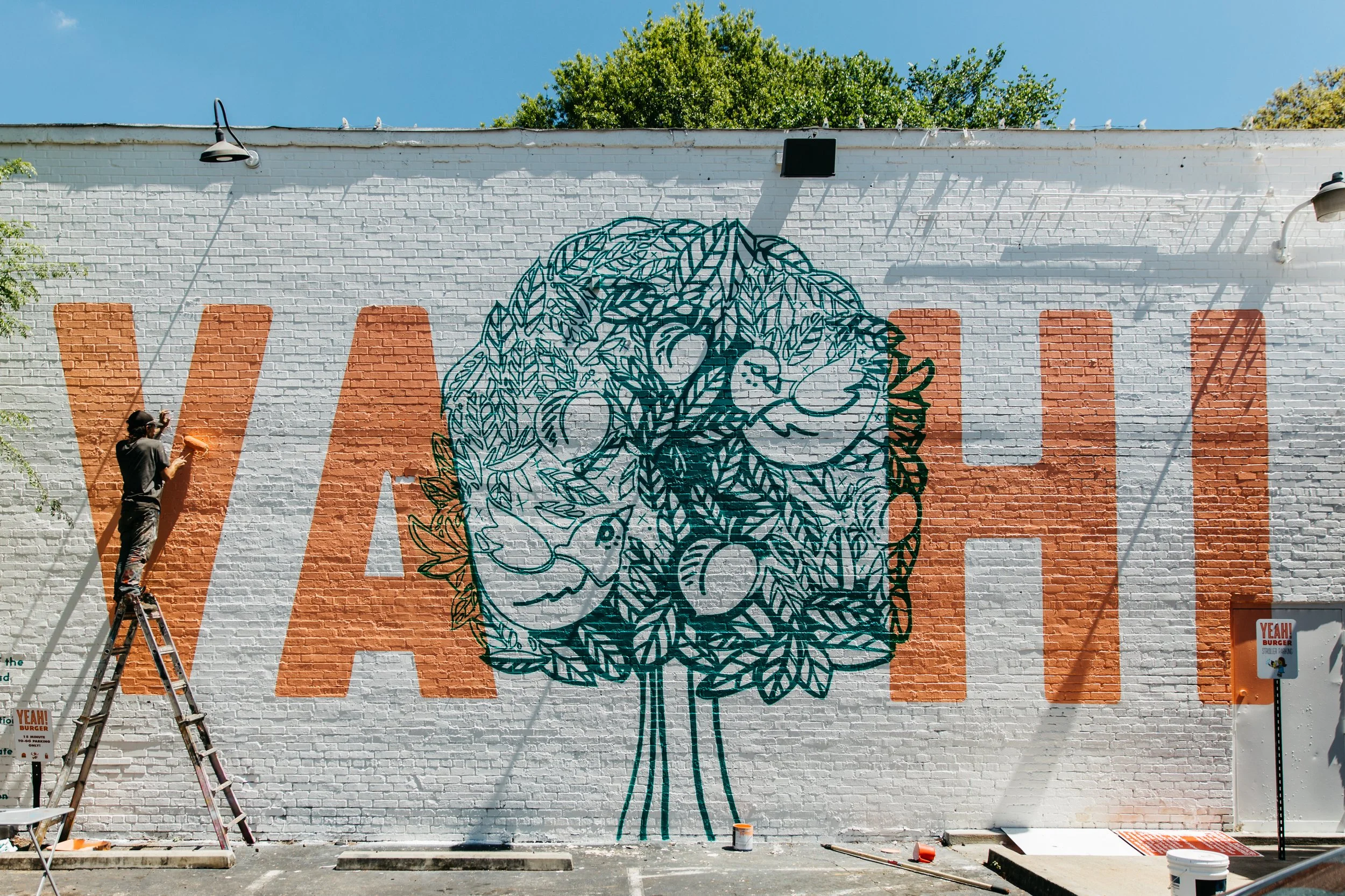 Artist painting a mural of a lion's face on a white brick wall with orange accents, during daytime.