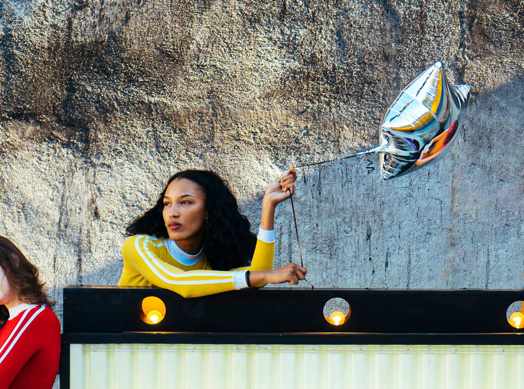 A woman with curly black hair, wearing a yellow top with white stripes on the sleeves, is sitting at a DJ booth against a textured gray wall. She is holding a silver balloon shaped like a star, attached to a thin string, and appears to be in a festiv