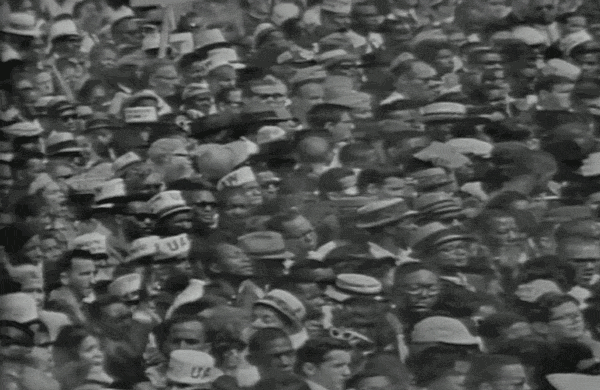 A large crowd of people gathered outdoors, many wearing hats and sunglasses.