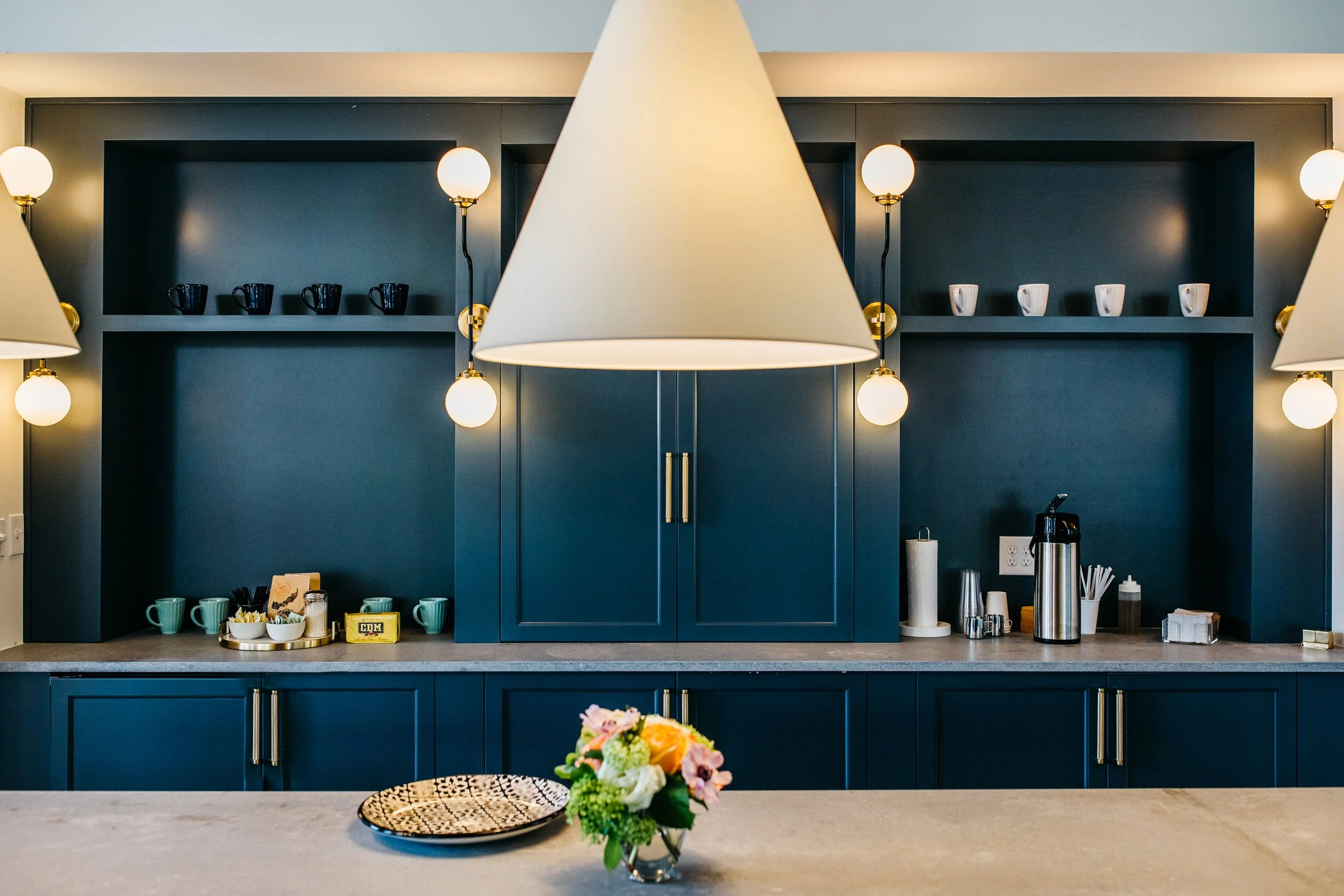 Modern kitchen with dark blue cabinets, hanging lights, and a marble countertop. Items on the counter include a flower vase, cups, and kitchen essentials.