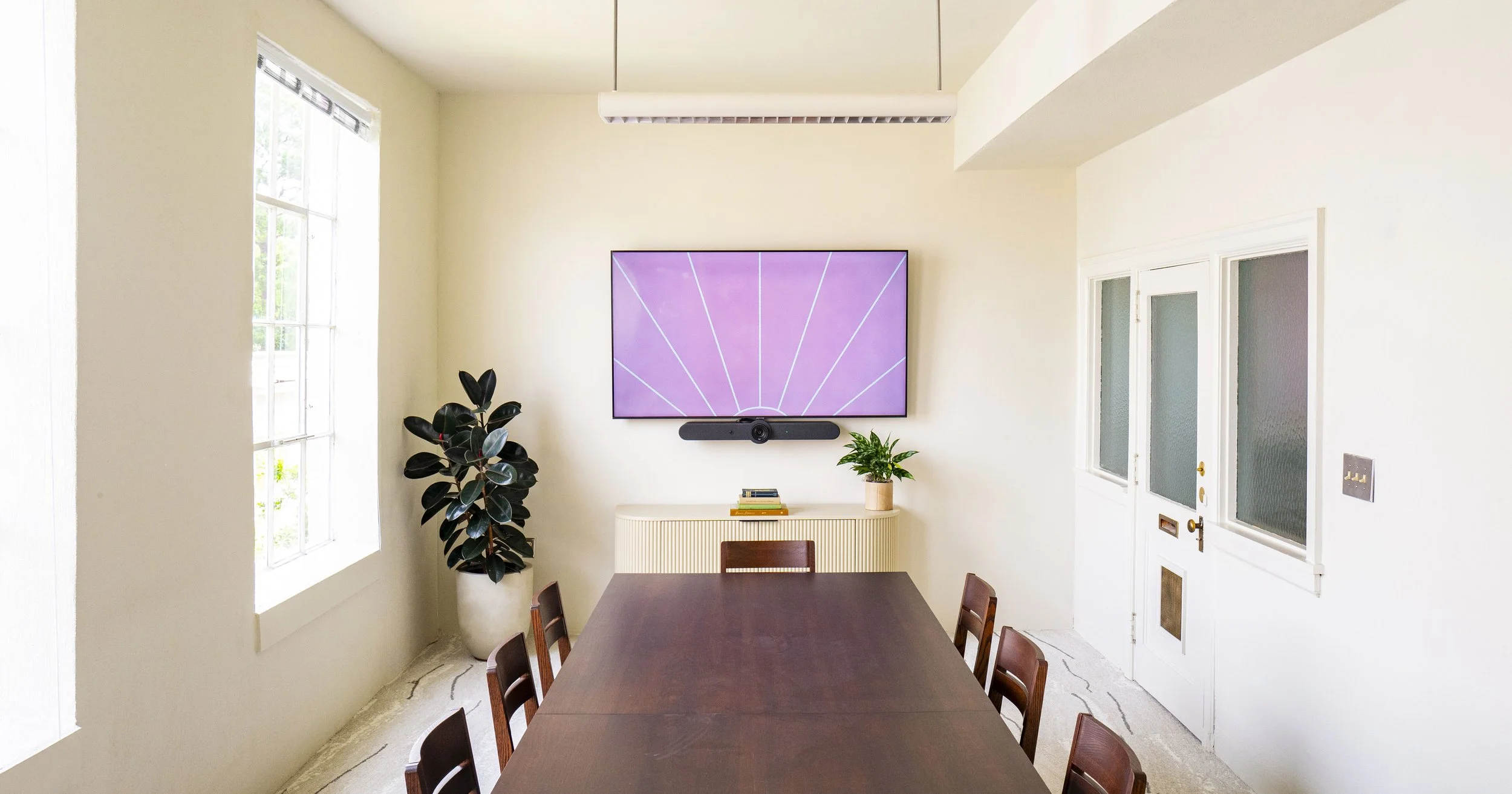 A dining room with a large wooden table, chairs, a wall-mounted flat-screen TV displaying a pink background with white lines, a soundbar underneath, a sideboard with books and a potted plant, a window on the left, and a door on the right.