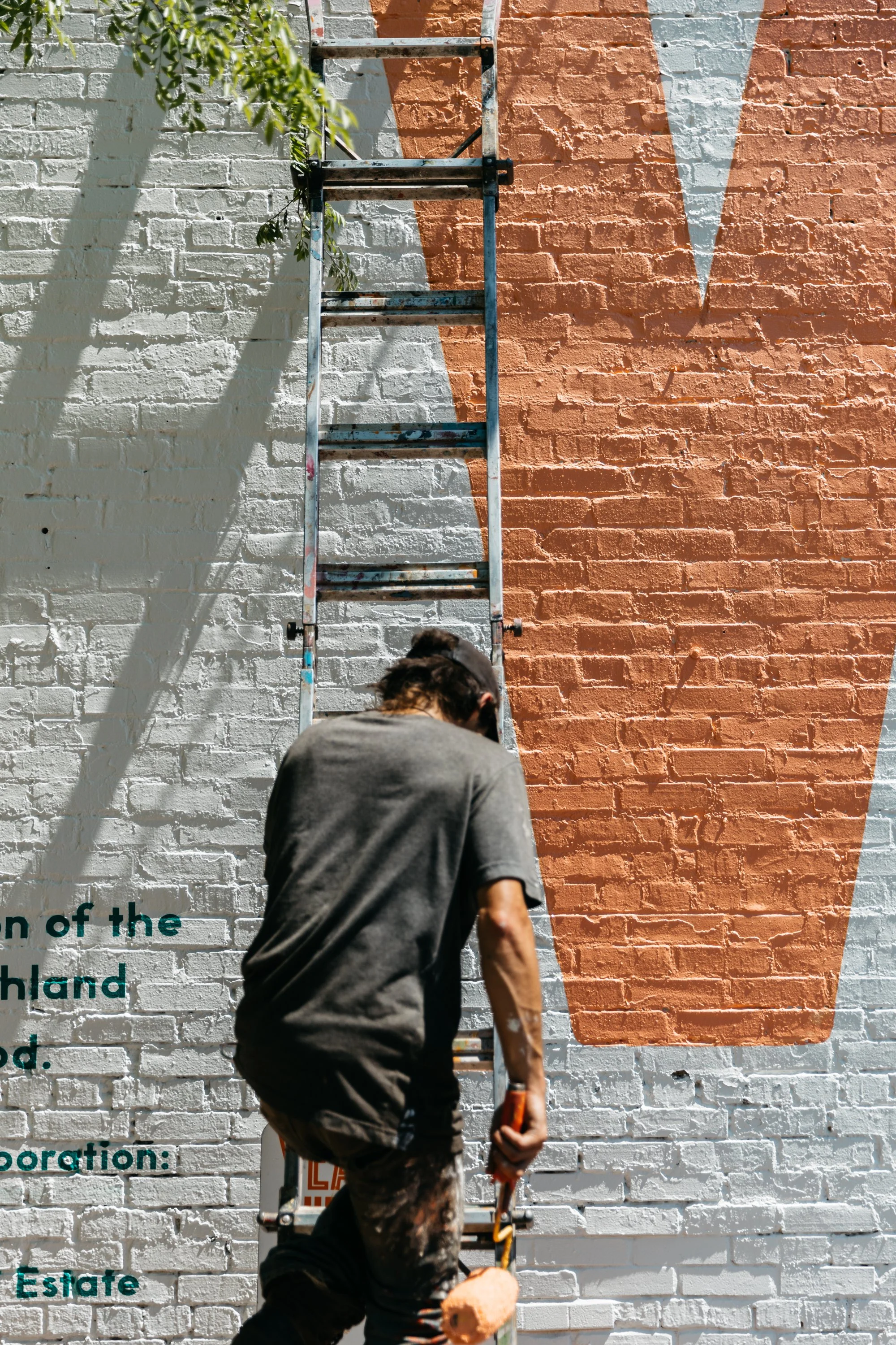 Person painting a mural on a brick wall with orange and white sections. The person is on a ladder, holding a paint roller, wearing dark clothing, and has dark curly hair.