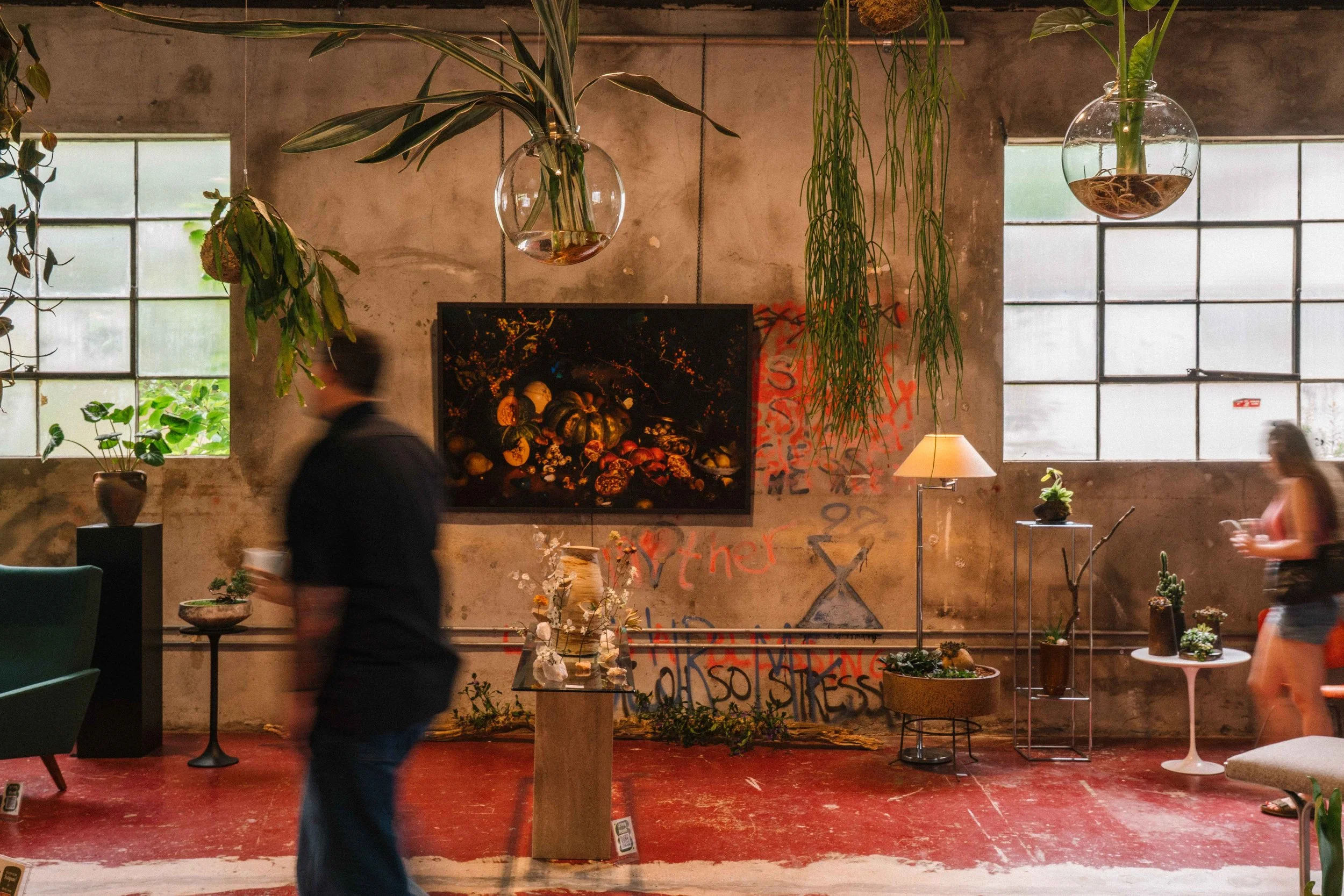 Interior of a room with large industrial-style windows, hanging glass planters with plants, a TV screen with artwork, and various plants and furniture, with two blurred people walking past.