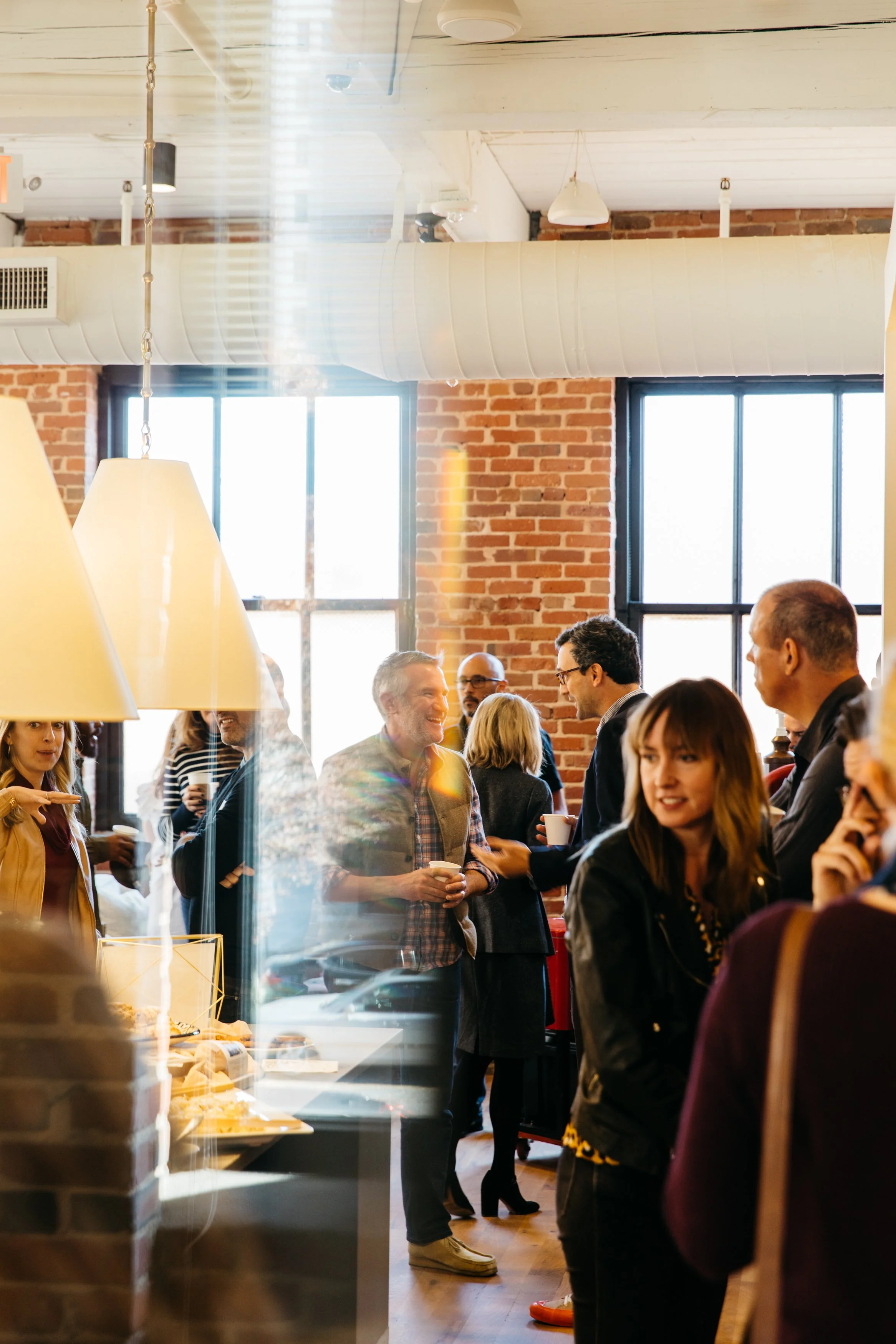 People socializing at a cafe or bakery with exposed brick walls and large windows, some holding cups, in a lively and warm atmosphere.