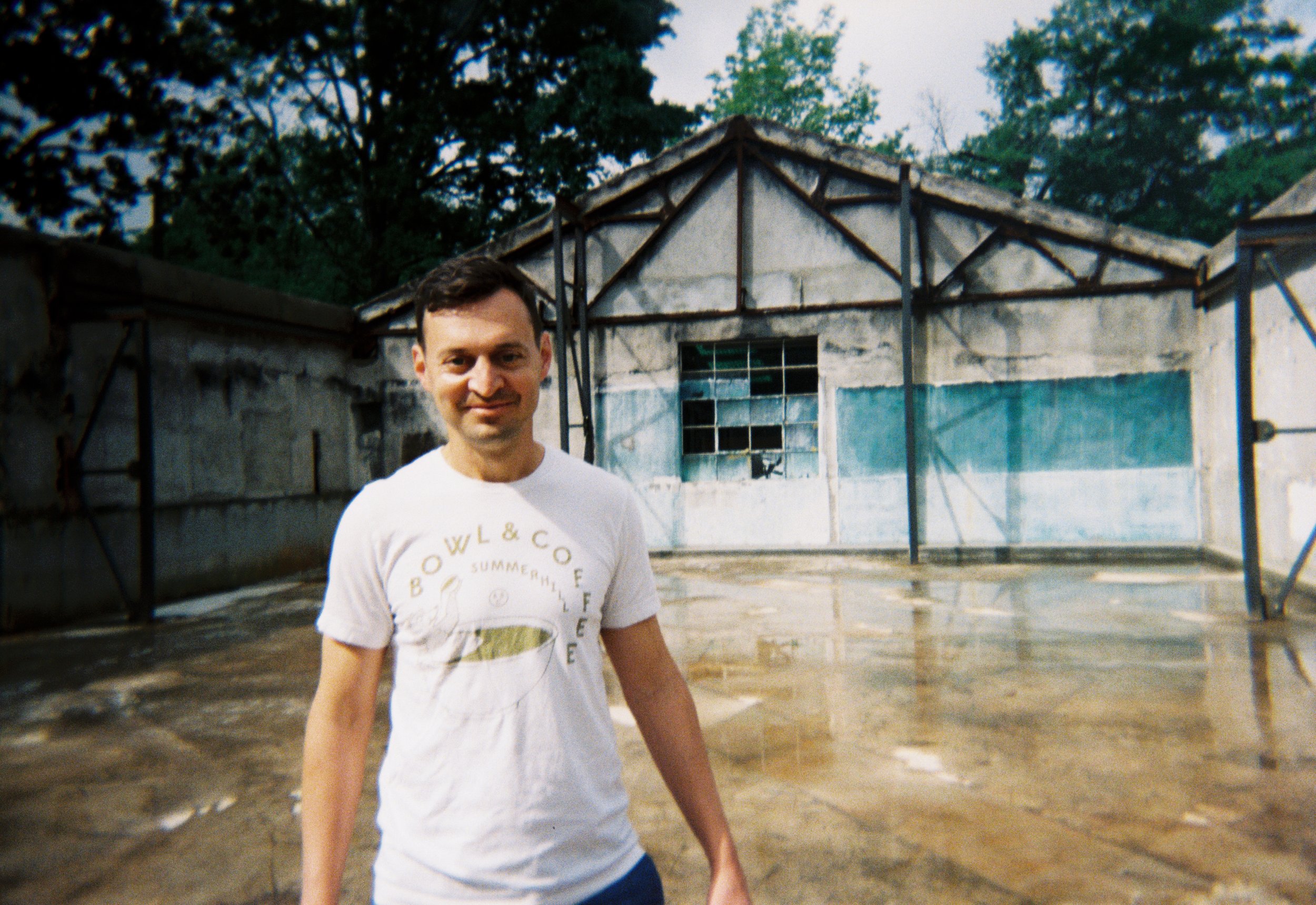 A smiling man wearing a white t-shirt stands outdoors in front of a partially constructed building with scaffolding, surrounded by trees.