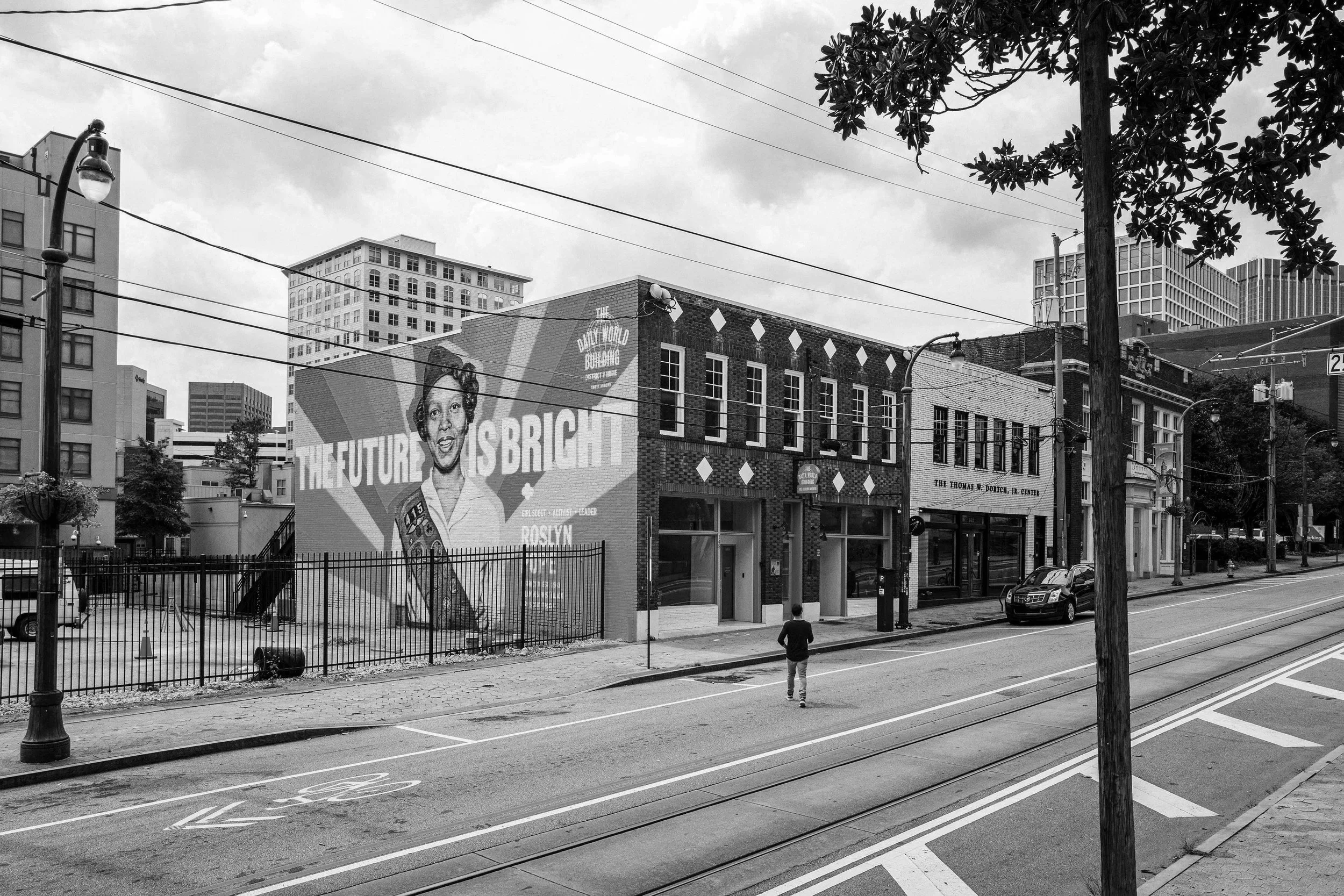 A city street scene with a large billboard featuring a portrait of a woman and a message that says 'The Future is Bright'. The street has some parked cars, a pedestrian walking, streetlights, power lines, and trees in the background with buildings visible.
