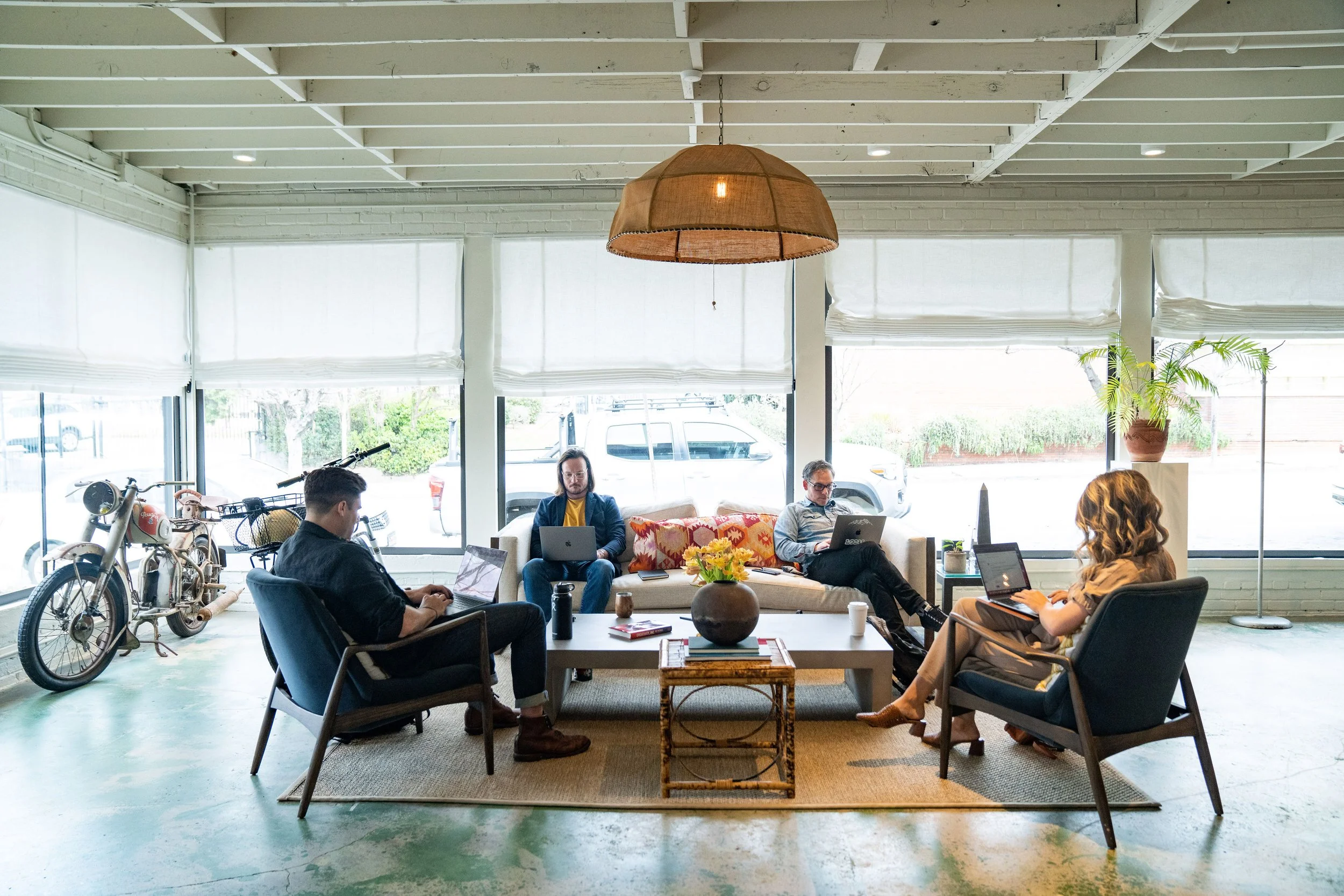 A lively coffee shop with large windows, white brick walls, and a high ceiling with exposed beams. Several people are seated, working on laptops, with bikes parked inside and a potted plant near the window. A wooden pendant light hangs from the ceiling.